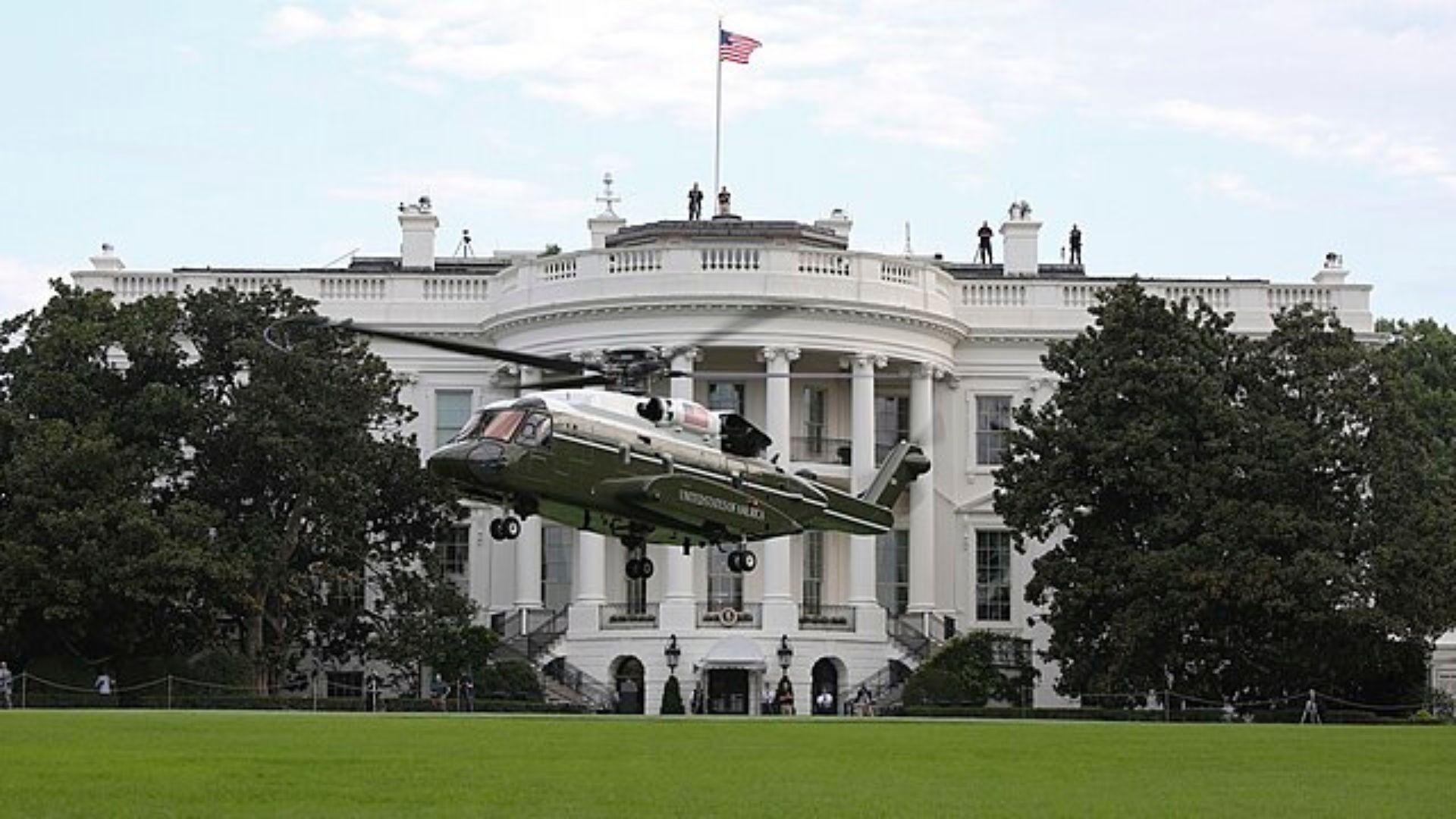 The VH-92 helicopter preparing to land on the lawn in front of the White House. The American flag is flying on top of the White House.