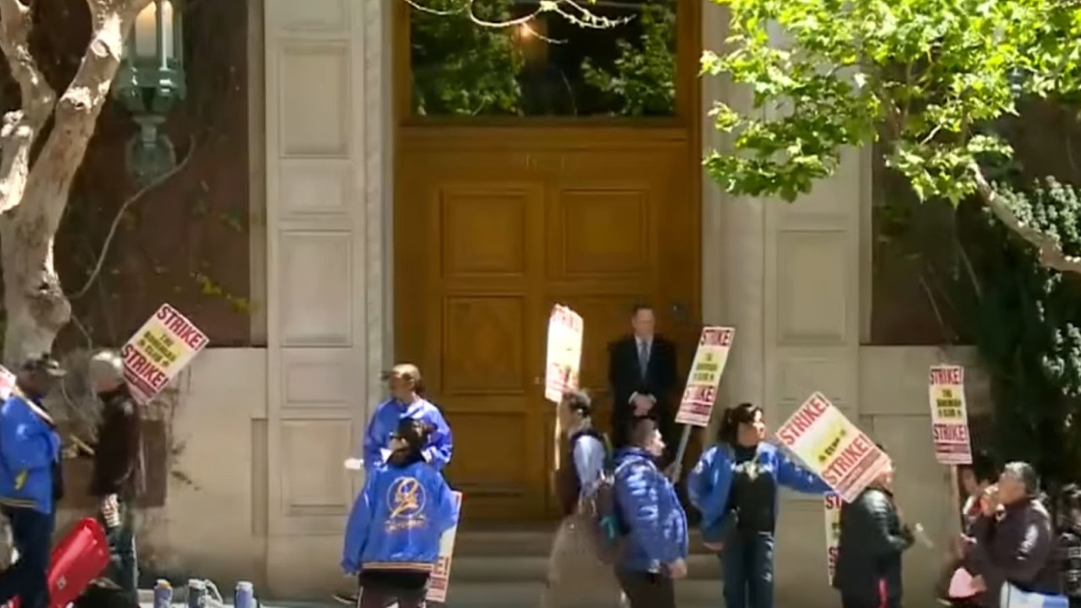 The Bohemian Club workers are going on strike. They are holding placards on wooden sticks in the air that say “STRIKE!” in red capital letters.