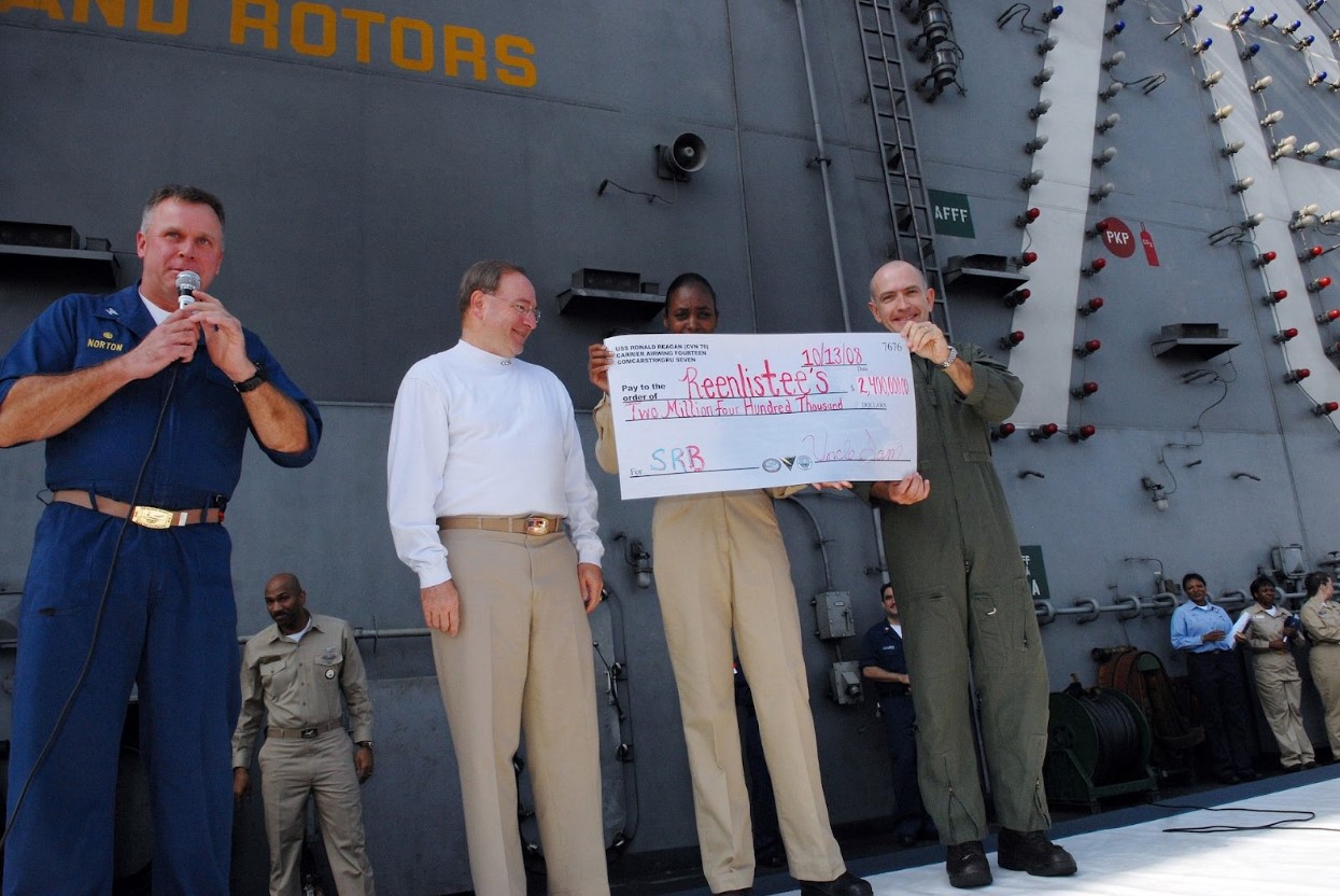 A few people standing in Navy uniform standing with a mock-up check addressed to re-enlistees