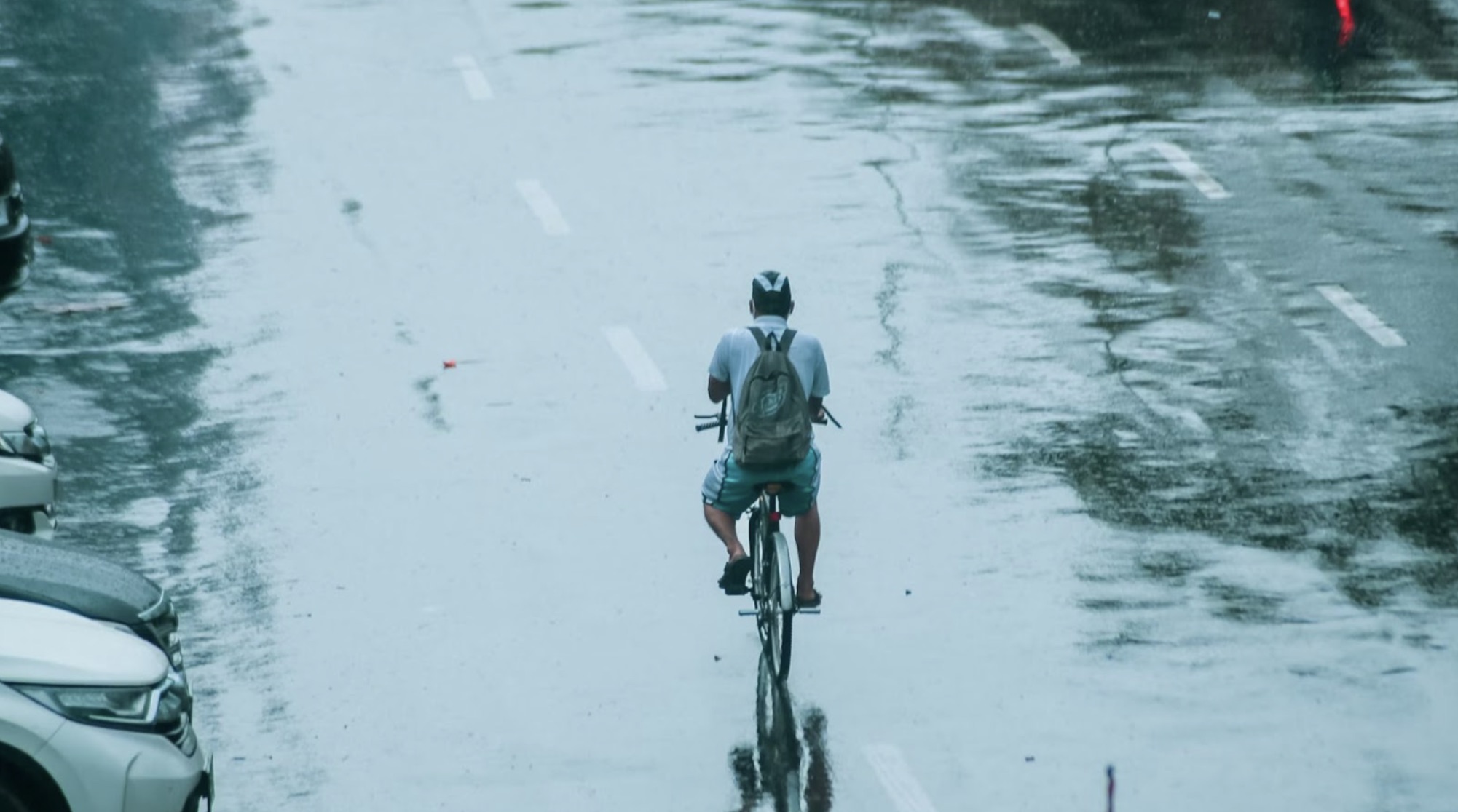 Flooding on the road in Jakarta. A person on the bike is riding through the floods. Cars are parked on the side of the road.