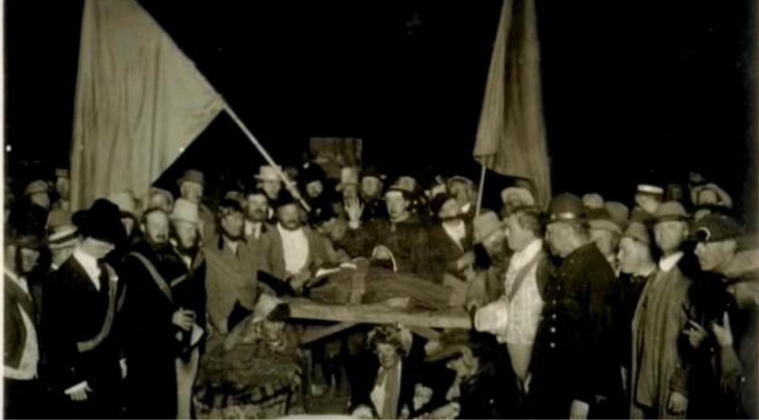 This is a black-and-white photo of Bohemian Grove. There is a crowd of people, two of whom are holding large flags on flagpoles.