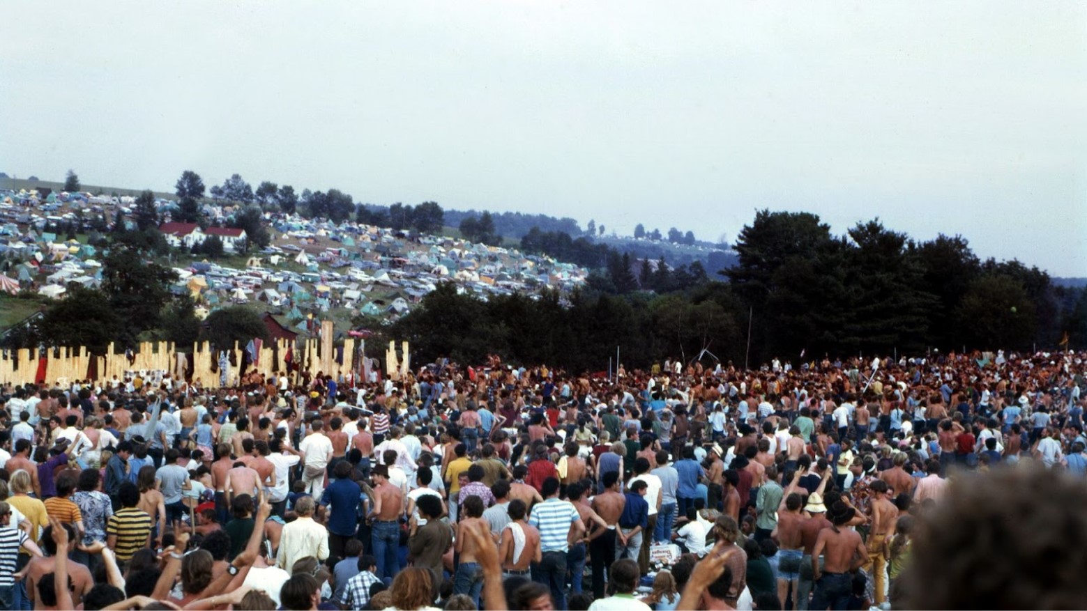 A large crowd at Woodstock, with tents and a home seen in the distance.