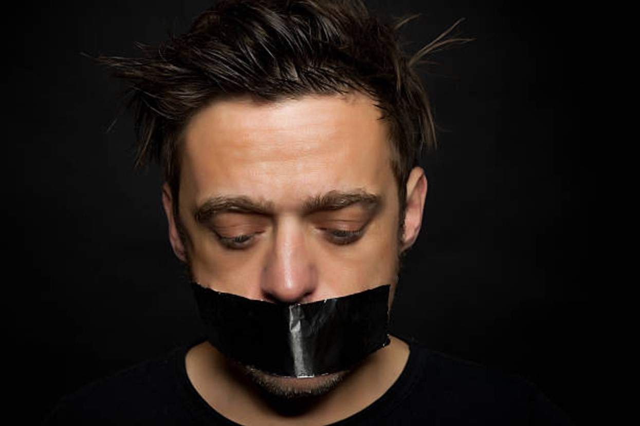 A man against a black background with a piece of black tape across his mouth