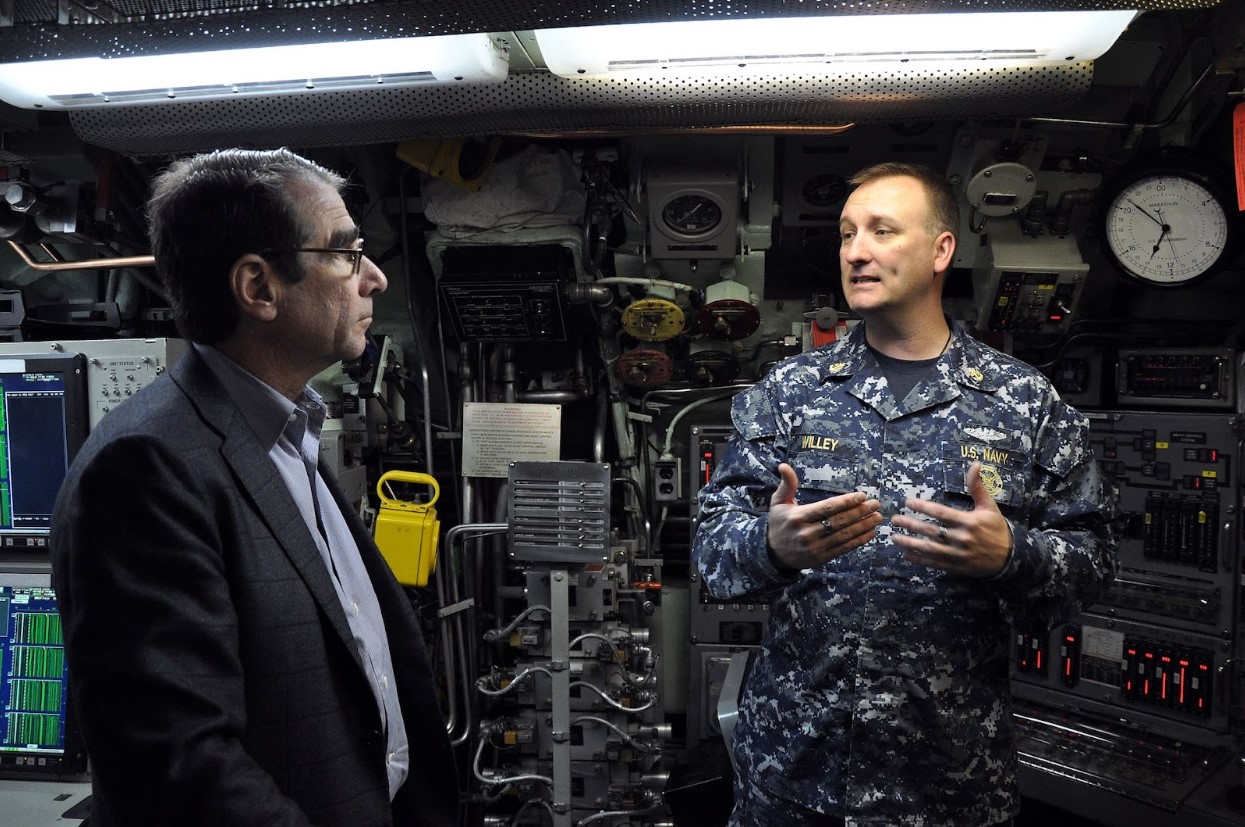 A man in Navy uniform talking to a man in a suit while standing in front of a control panel