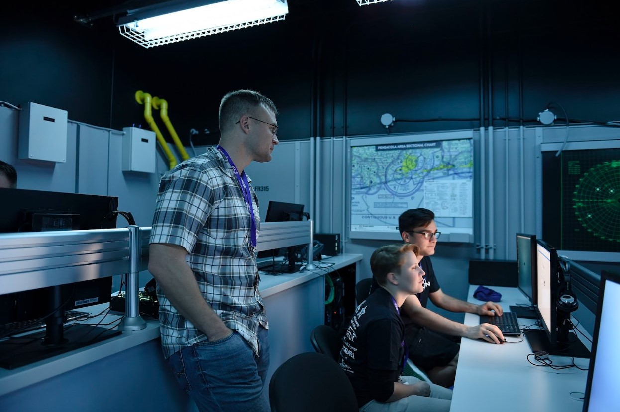 Two people working on a computer on a desk while a person stands behind them