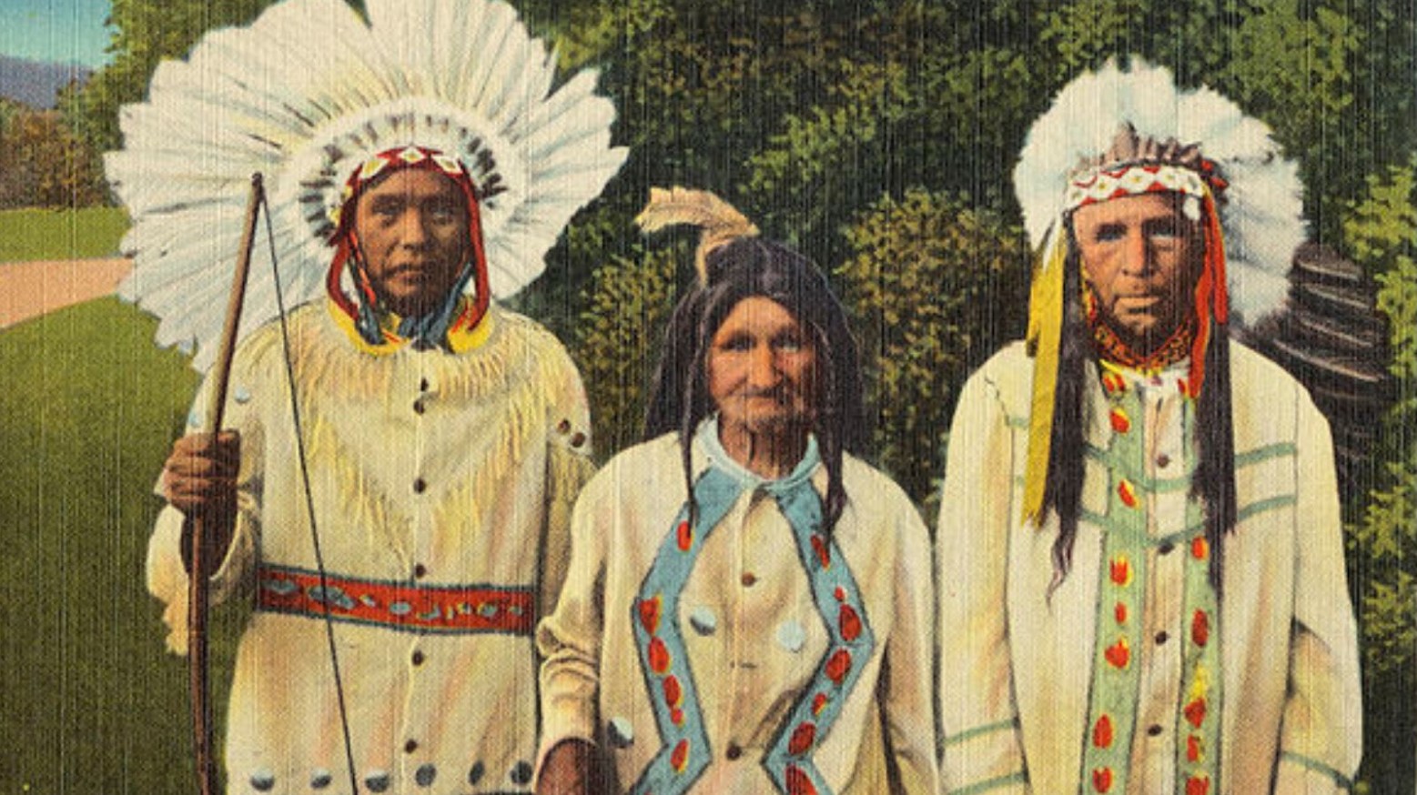 Three Cherokee Indians are standing in front of green bushes, wearing traditional Indian headdresses and clothes.