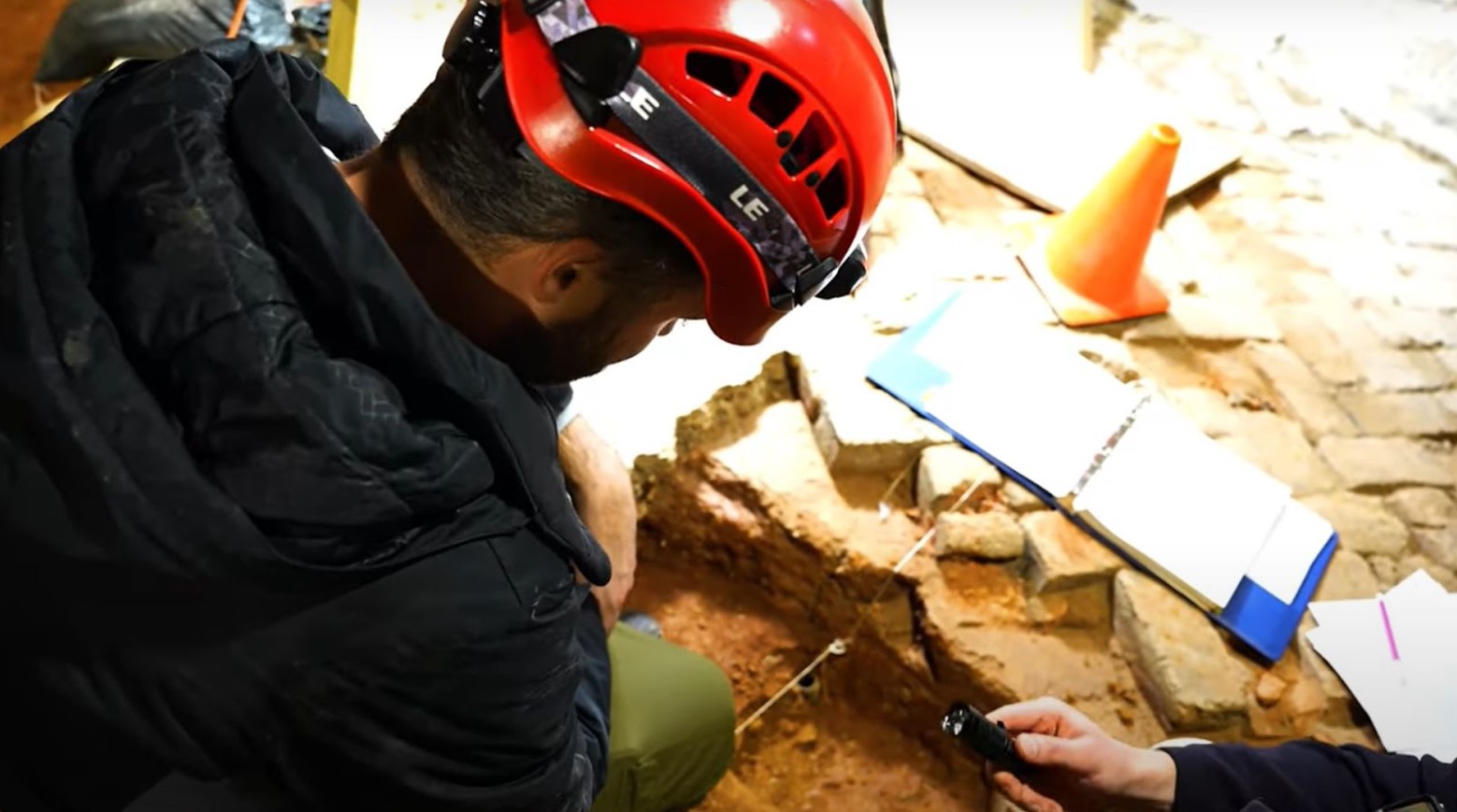 One of the bottles poking up from underneath the soil. An archaeologist, wearing a hard hat, is looking at the bottle with a torch. An open blue notepad and an orange cone are in front of him.