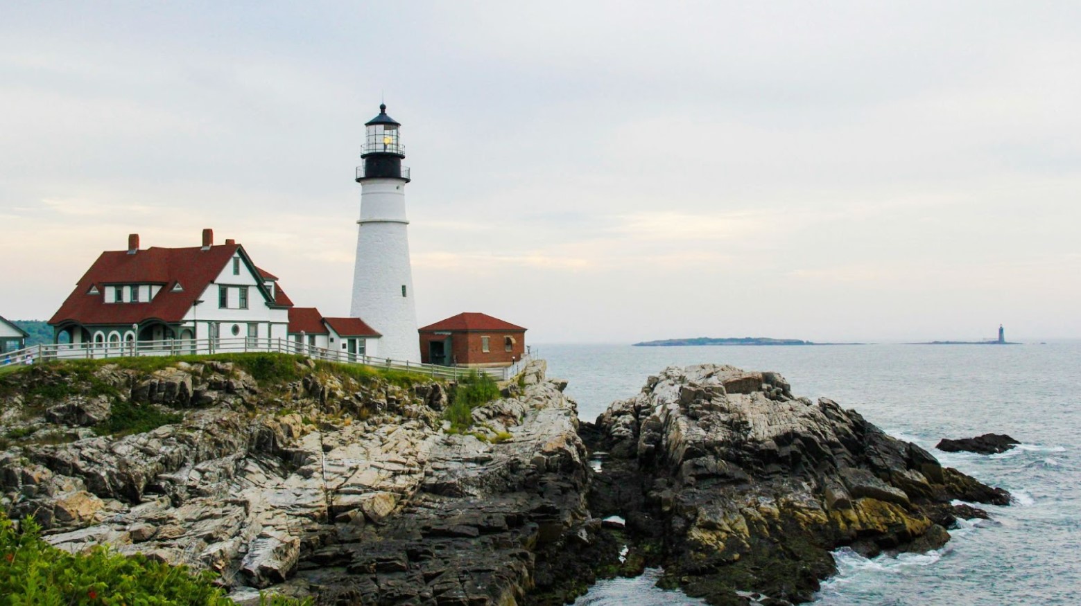 A building and lighthouse seen on a rocky coastline of Maine in the daytime.