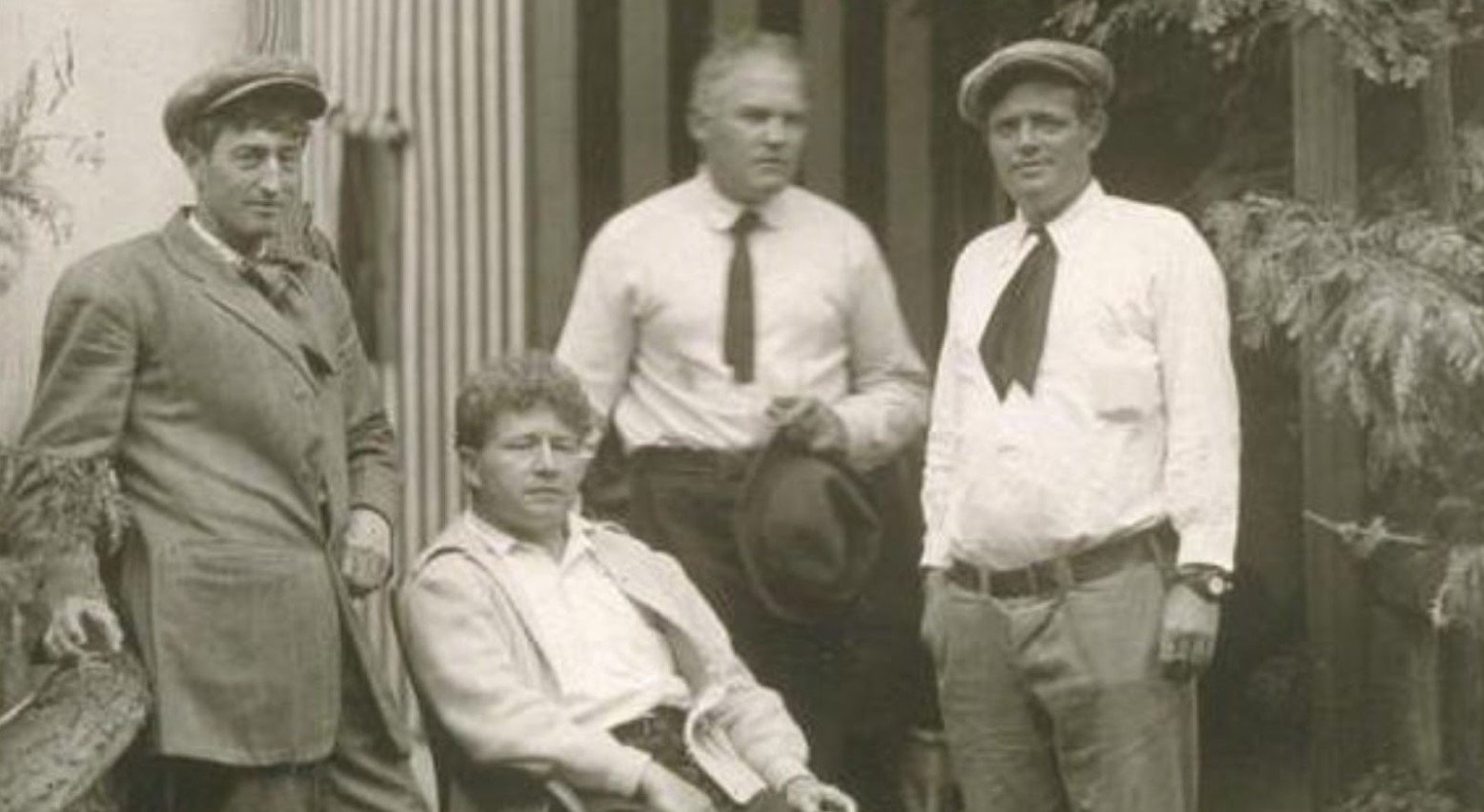 This is a black-and-white photo of four men at Bohemian Grove. They are wearing white shirts and trousers. One is wearing a coat, and they each have hats.