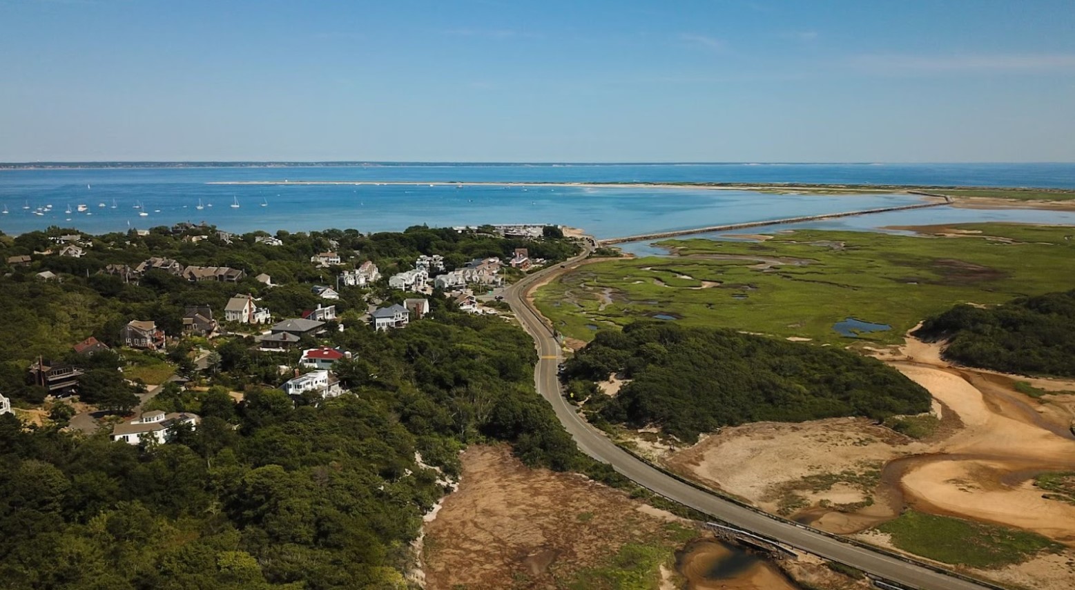 An aerial view of part of Massachusetts. A body of water is in the distance, and a road runs through the town. Houses are on the left of the image, and fields and sand are on the right.