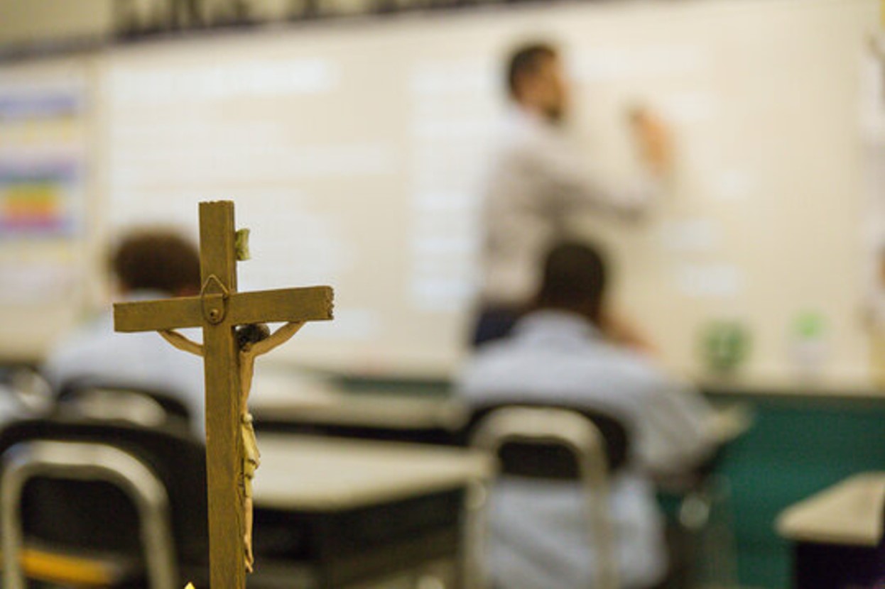 Close-up photograph of a Christian crucifix in front of a blurred background of a teacher in a classroom