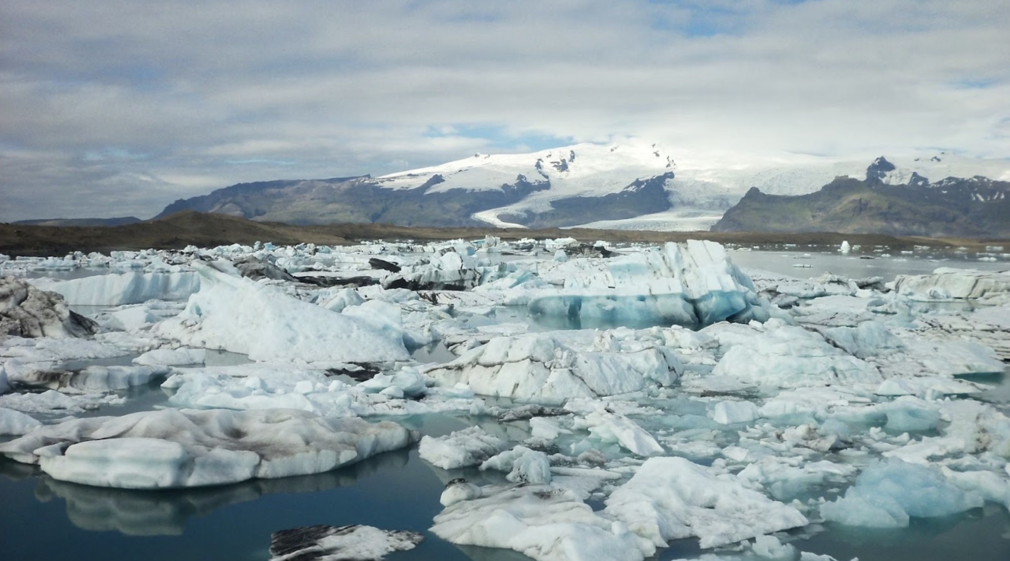 Melting polar ice caps. The ice caps are broken up and are in the water. Mountains with snow on top are in the background.