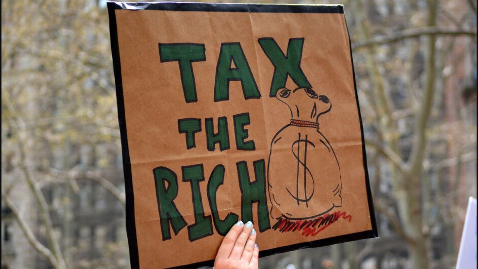 A hand holds up a cardboard sign that reads “Tax the Rich”