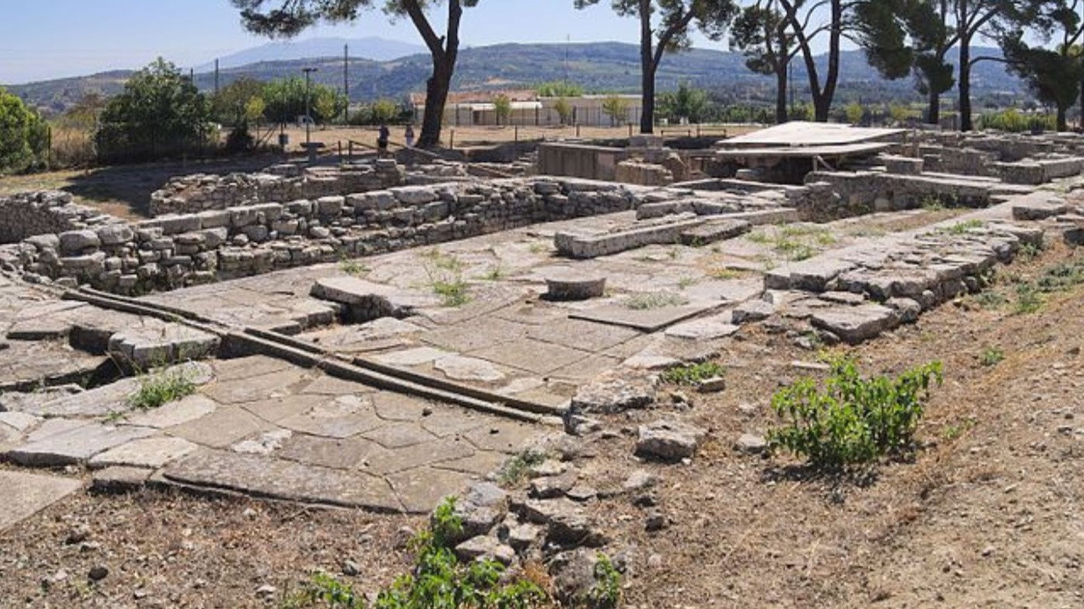 An ancient sanctuary at Tylissos in Crete. There are hills and trees in the background. The actual sanctuary consists of stones and rocks and is pretty empty.