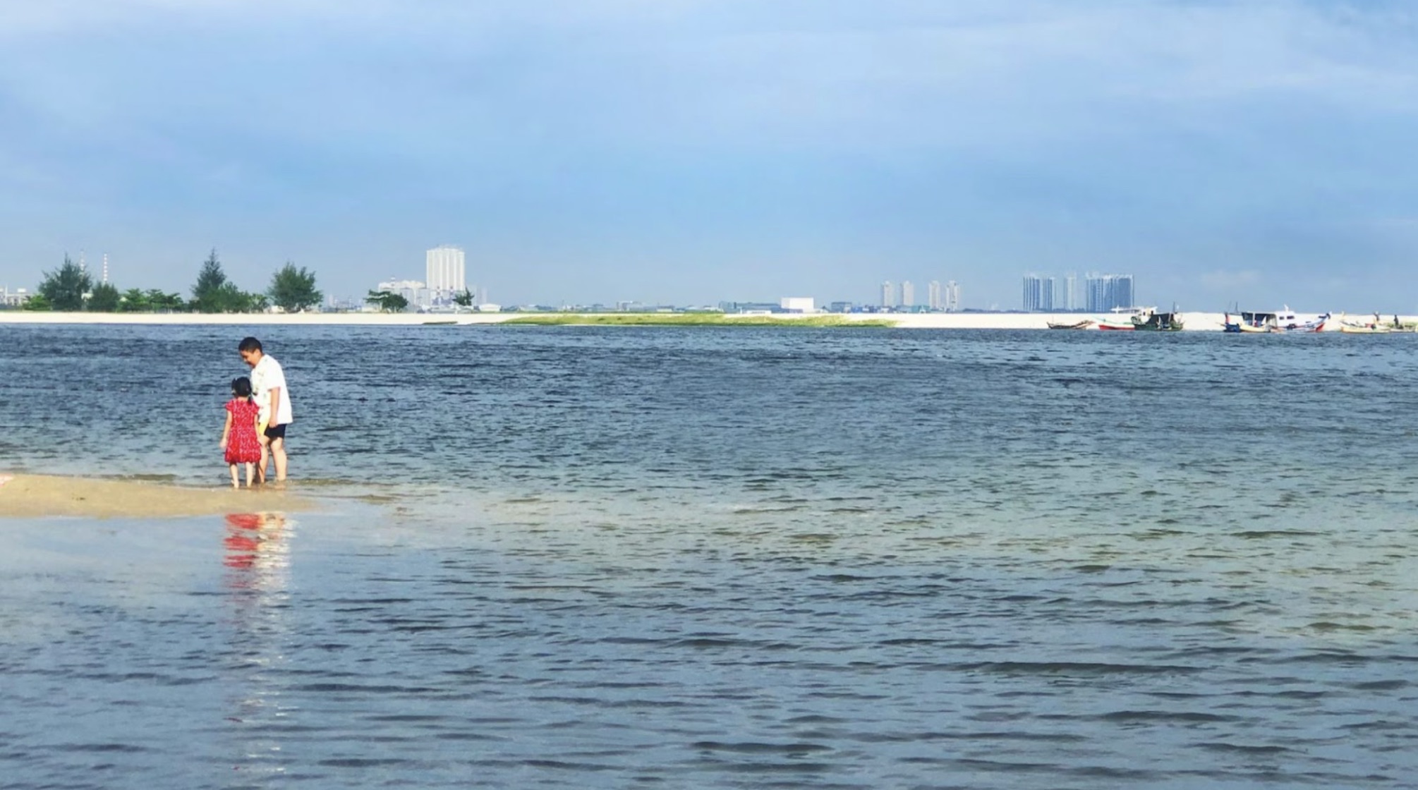 Two people standing on a small bit of land next to some water. Buildings are in the distance.