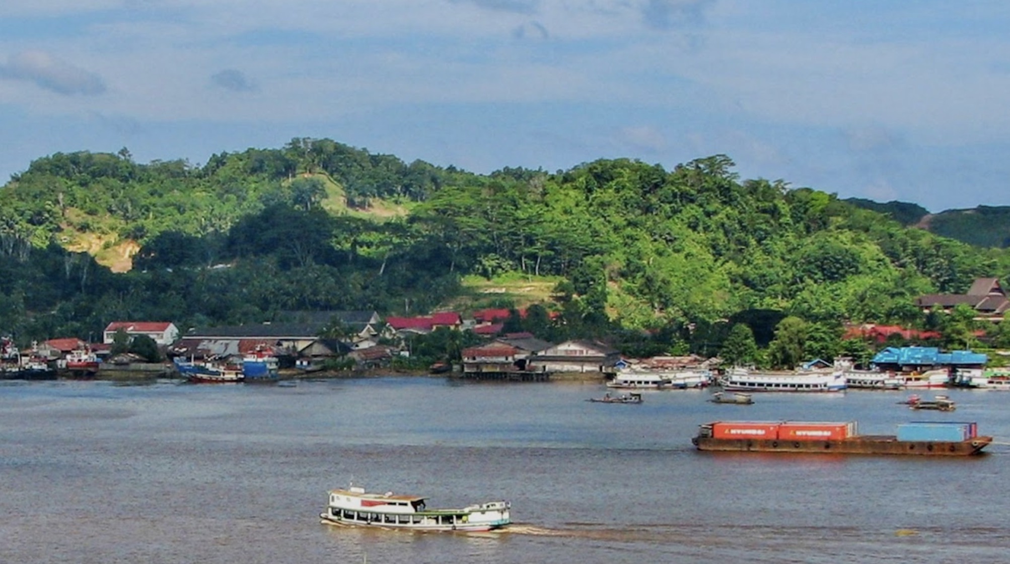 East Kalimantan in Indonesia. The area has boats on the water with houses next to the water. Behind them are green trees and fields.