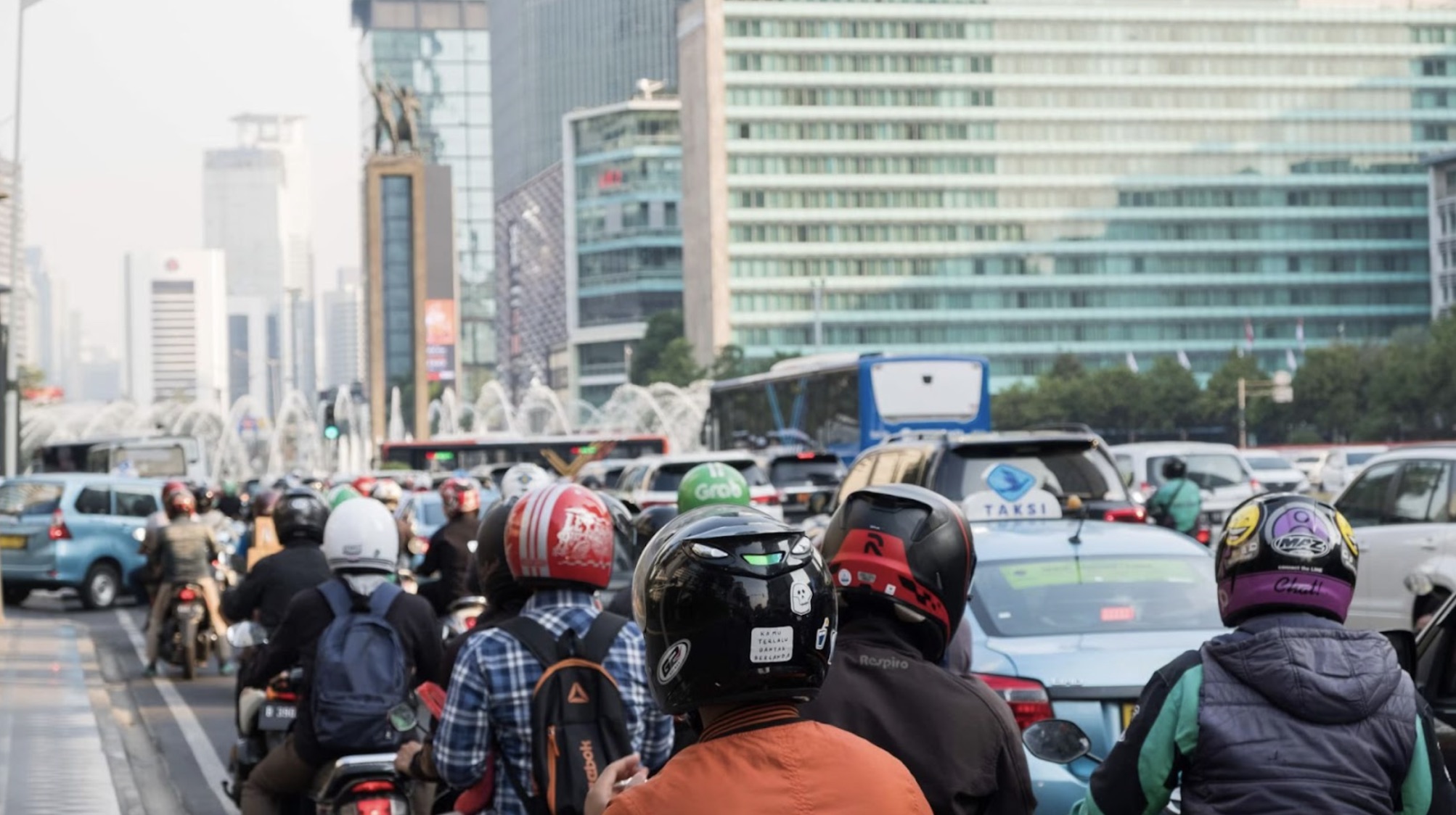A busy street in Jakarta. There are people on motorbikes and in cars and buses driving down the road, with high-rise buildings up ahead.