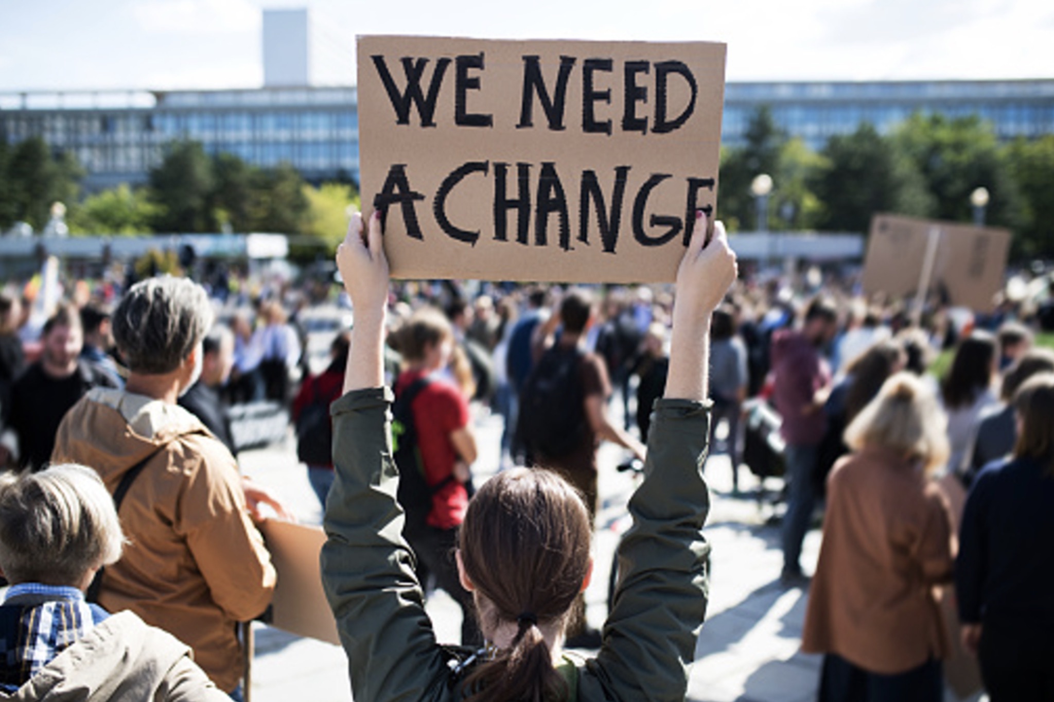Many people gather for a protest with one person holding a cardboard sign that reads “We Need A Change”