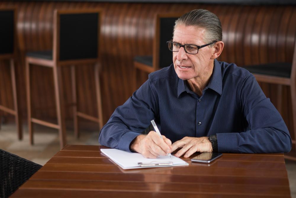 A photograph of an old man writing while being seated at a wooden table