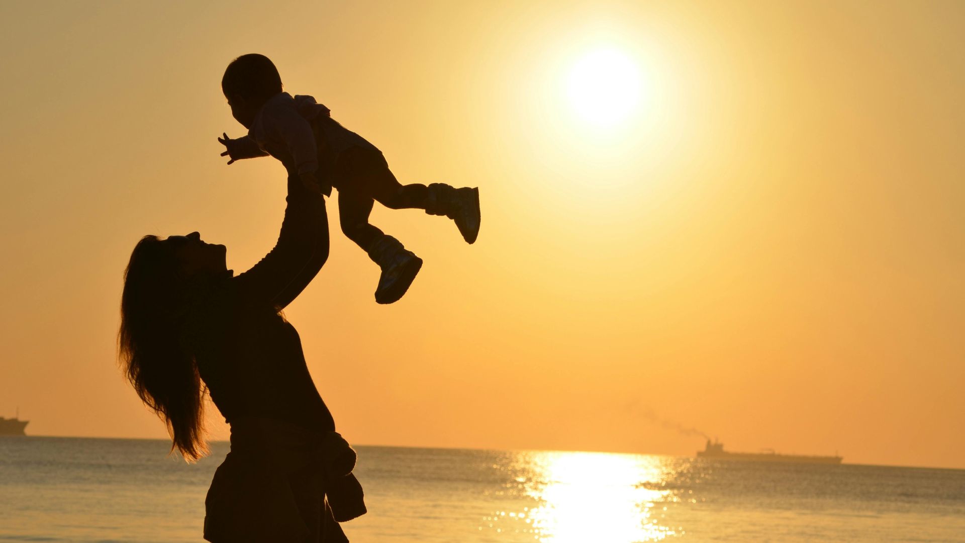 The silhouette of a mother raising her child above her head in front of the ocean during sunset.