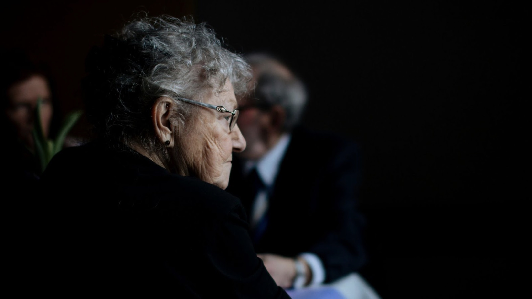 An elderly woman wearing glasses with people behind her.