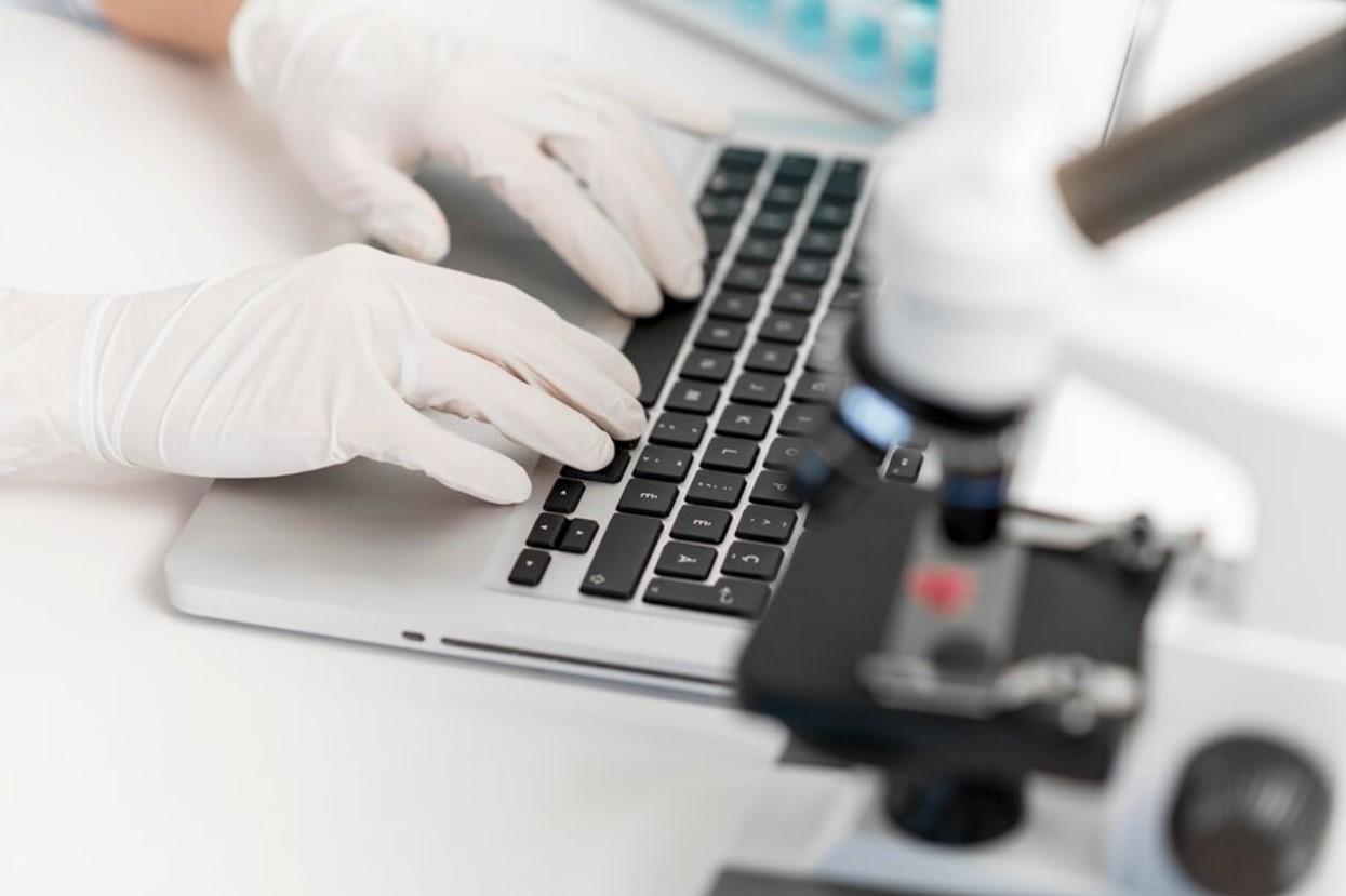 A faceless scientist wearing white gloves types on a laptop next to a microscope