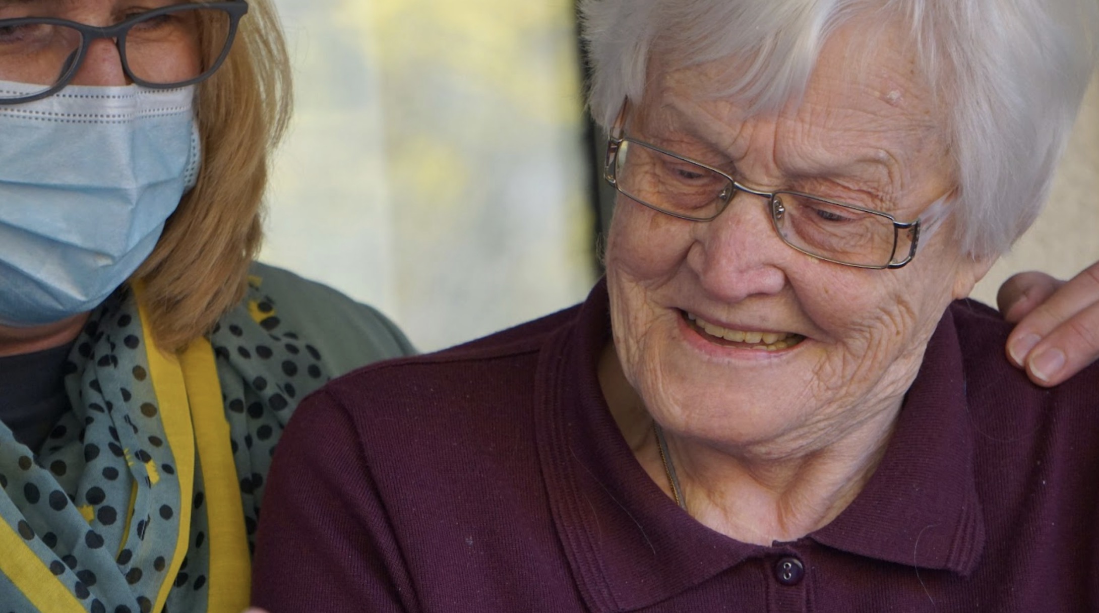 A woman wearing a mask helping an older senior citizen.