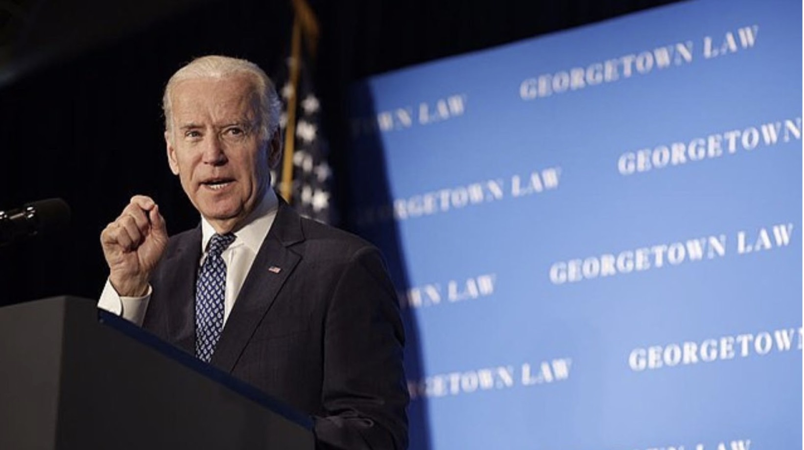 Joe Biden standing next to a lectern. Behind him is a blue background with “Georgetown Law” written in white.