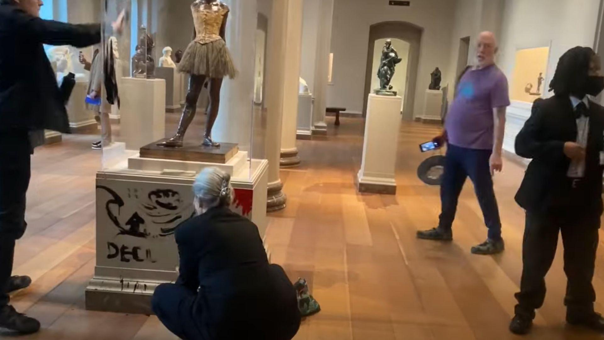 Joanna Smith and Timothy Martin defacing the Little Dancer Aged Fourteen statue. Martin is holding a water bottle in his hand with black paint inside. Martin covers the glass case in black paint, and Smith covers the base in red paint.
