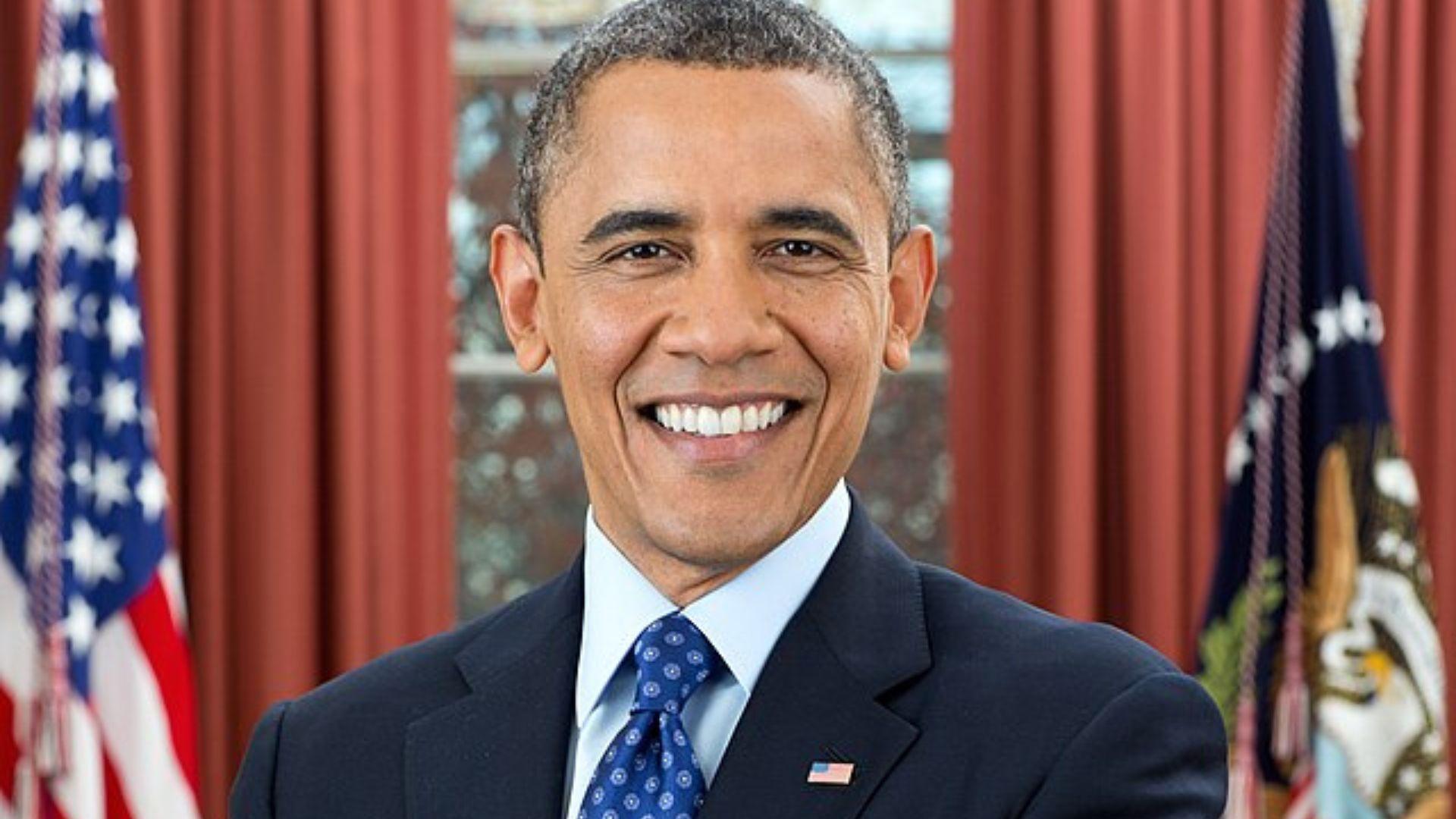 A portrait of Barack Obama. He is standing between two flags and has a pin on his suit of the US flag.