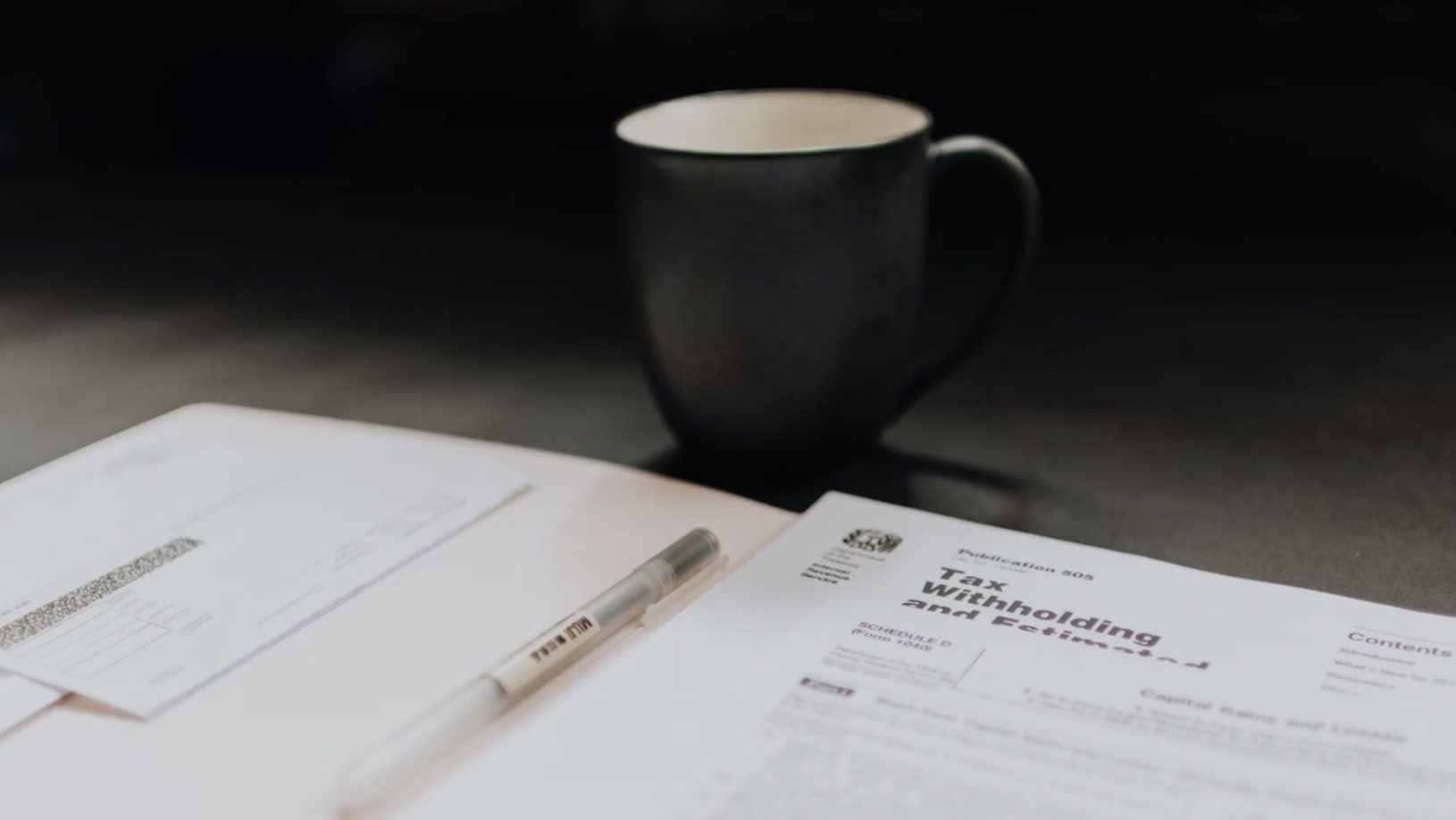 Some tax forms and a pen on a table next to a coffee mug.