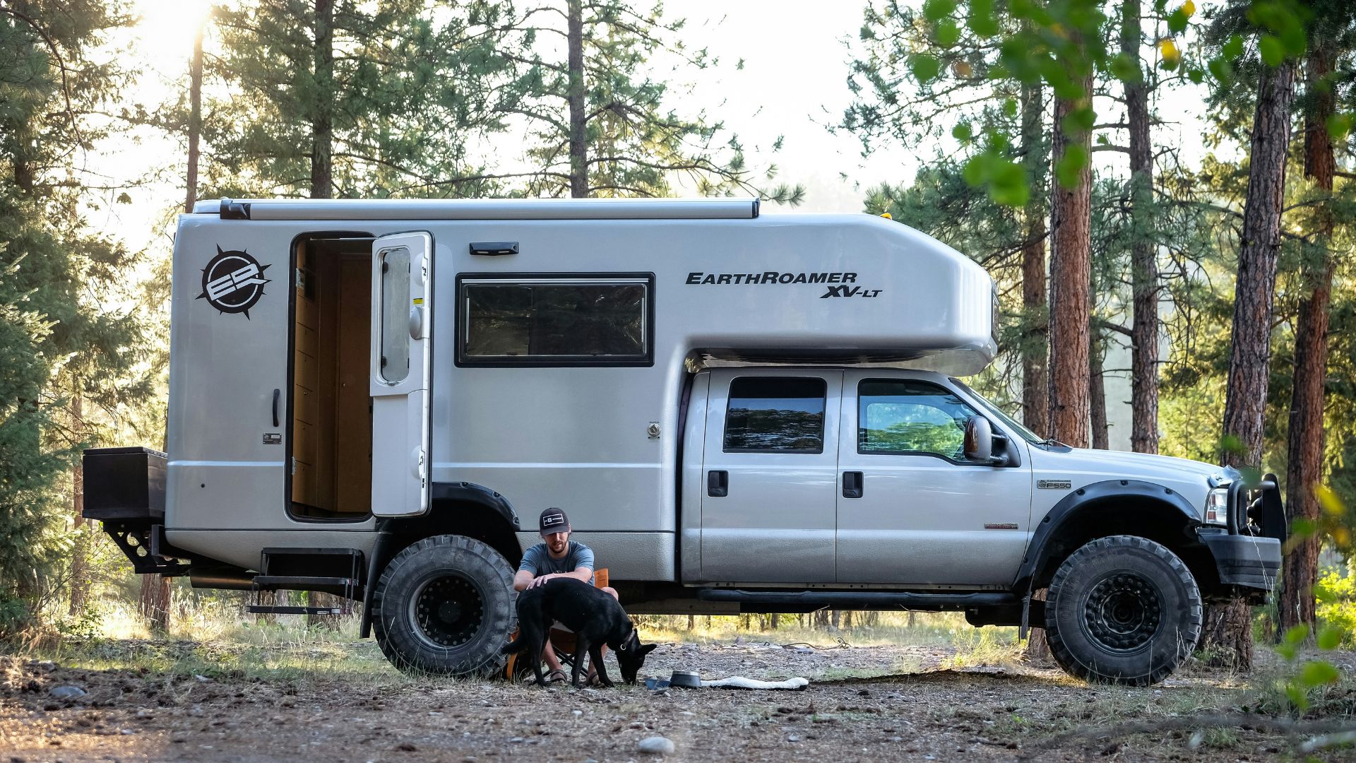 A man sitting outside with his dog in front of an RV in Montana in the daytime in the woods.