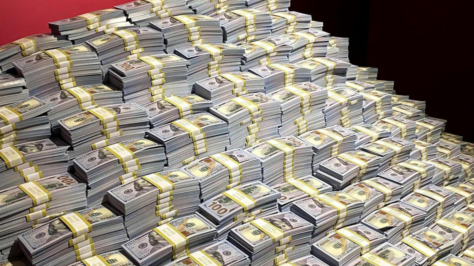 A mountain of $100 notes stacked up against a red wall.