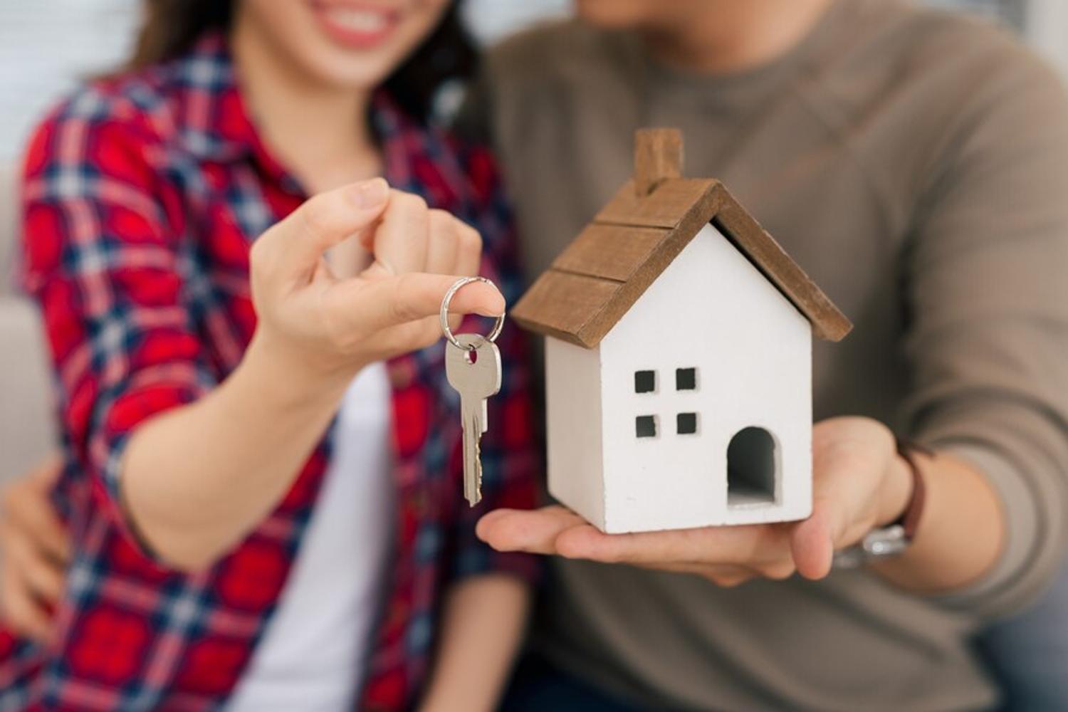 A young couple holds a small wooden home and a key depicting buying a new home