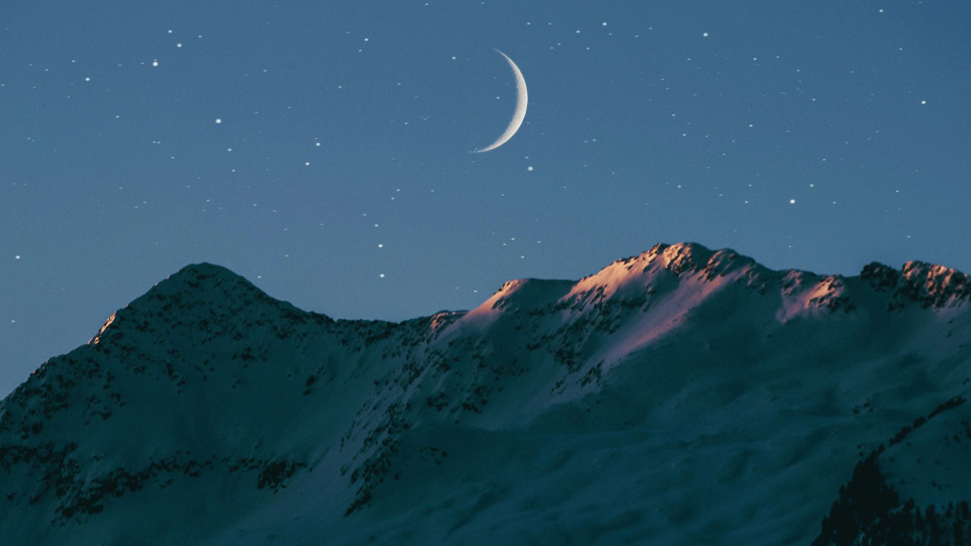 A crescent moon seen in a starry night sky above mountains.