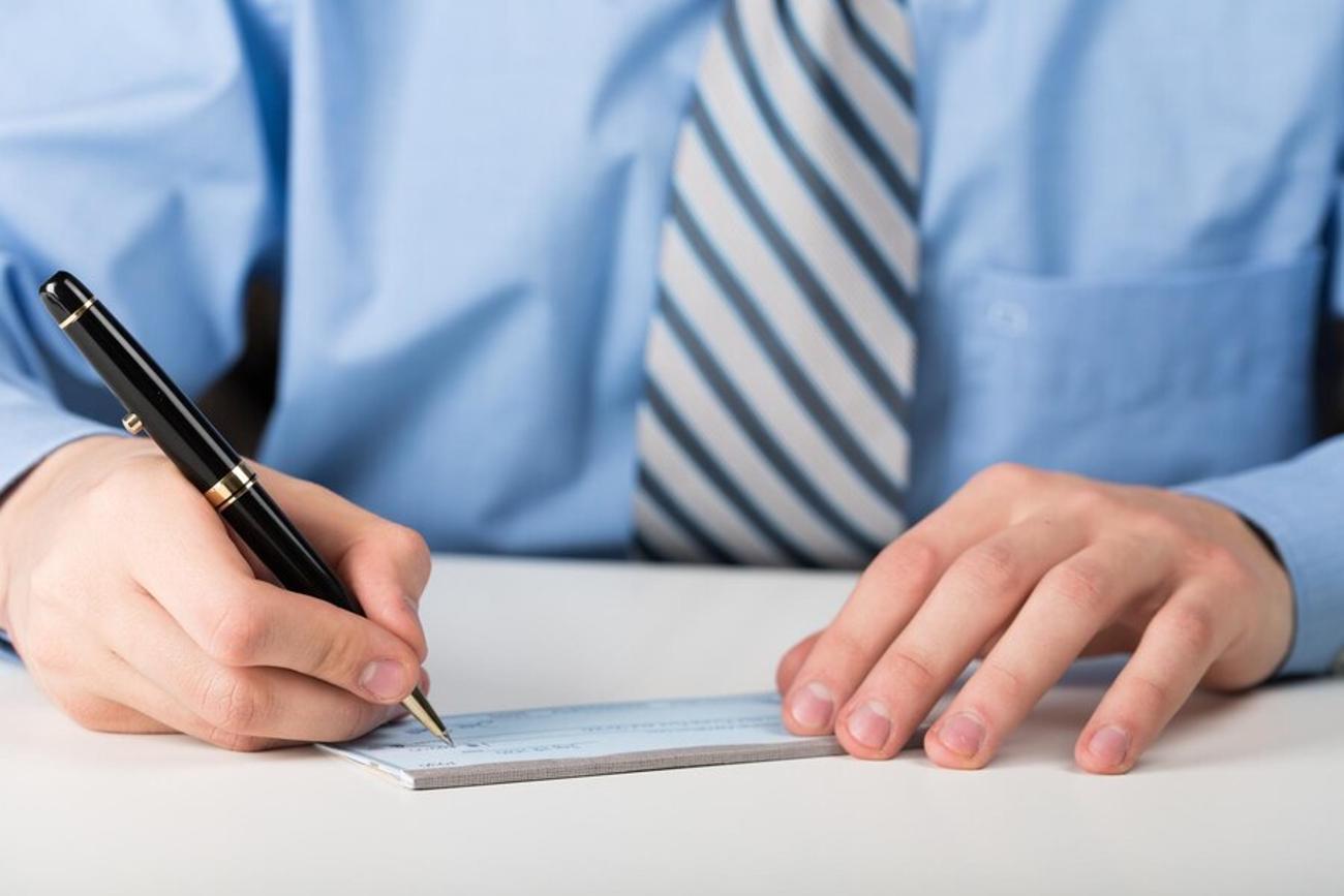 A mean in a blue shirt and tie writes a check with a black pen