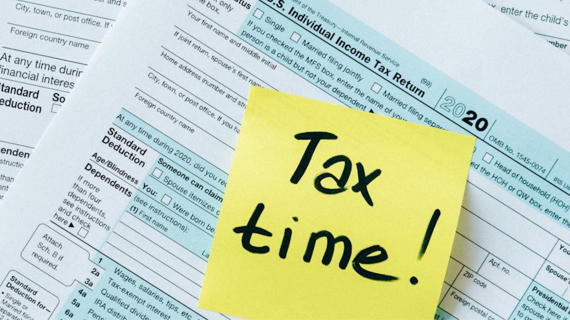 Tax return forms with a yellow sticky note on top that says “Tax time!” in black writing.
