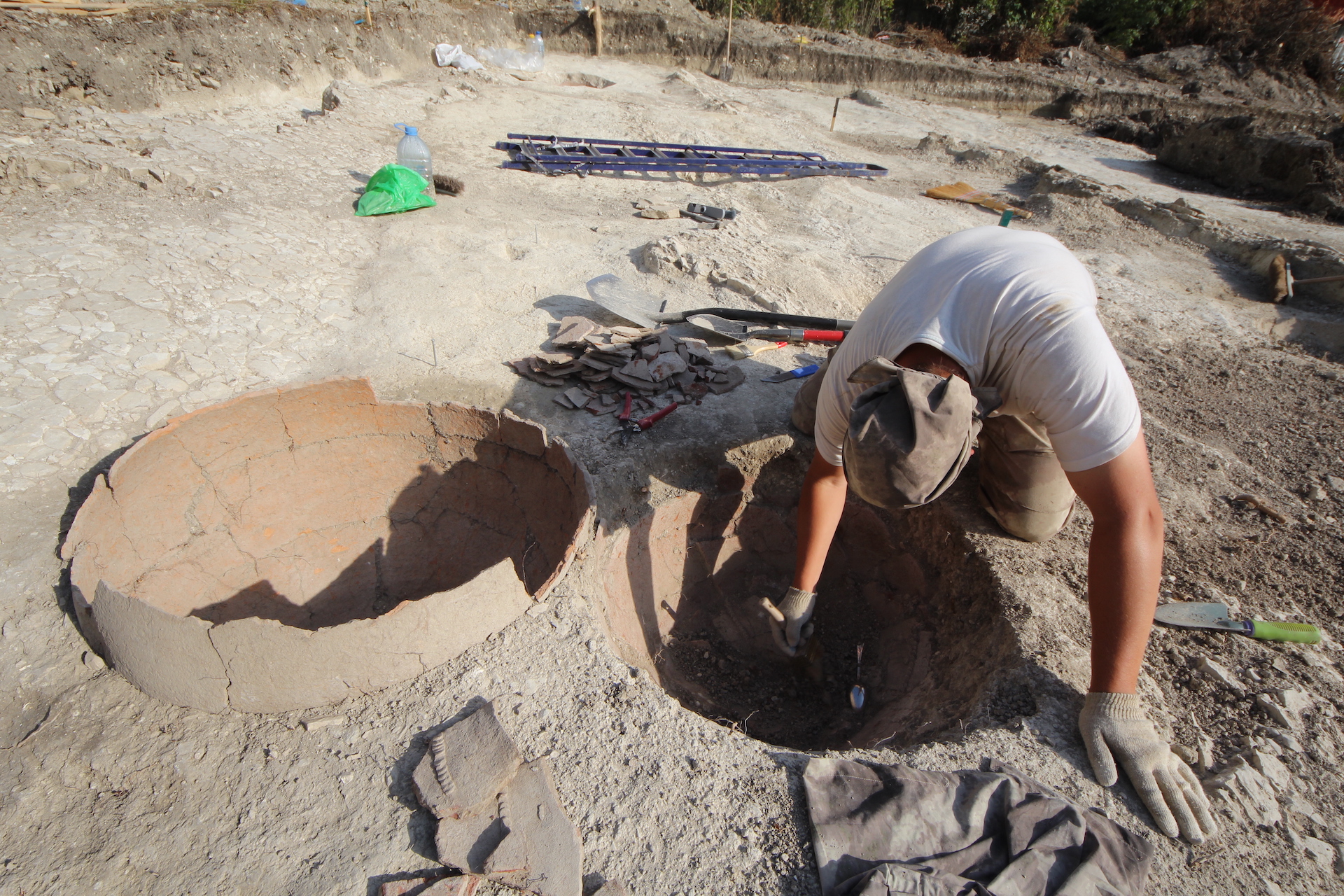 The archaeologist cleans the remains of a large ceramic vessel - pithos-from the ground with a brush. Another such vessel is installed nearby. In the background lie different tools.