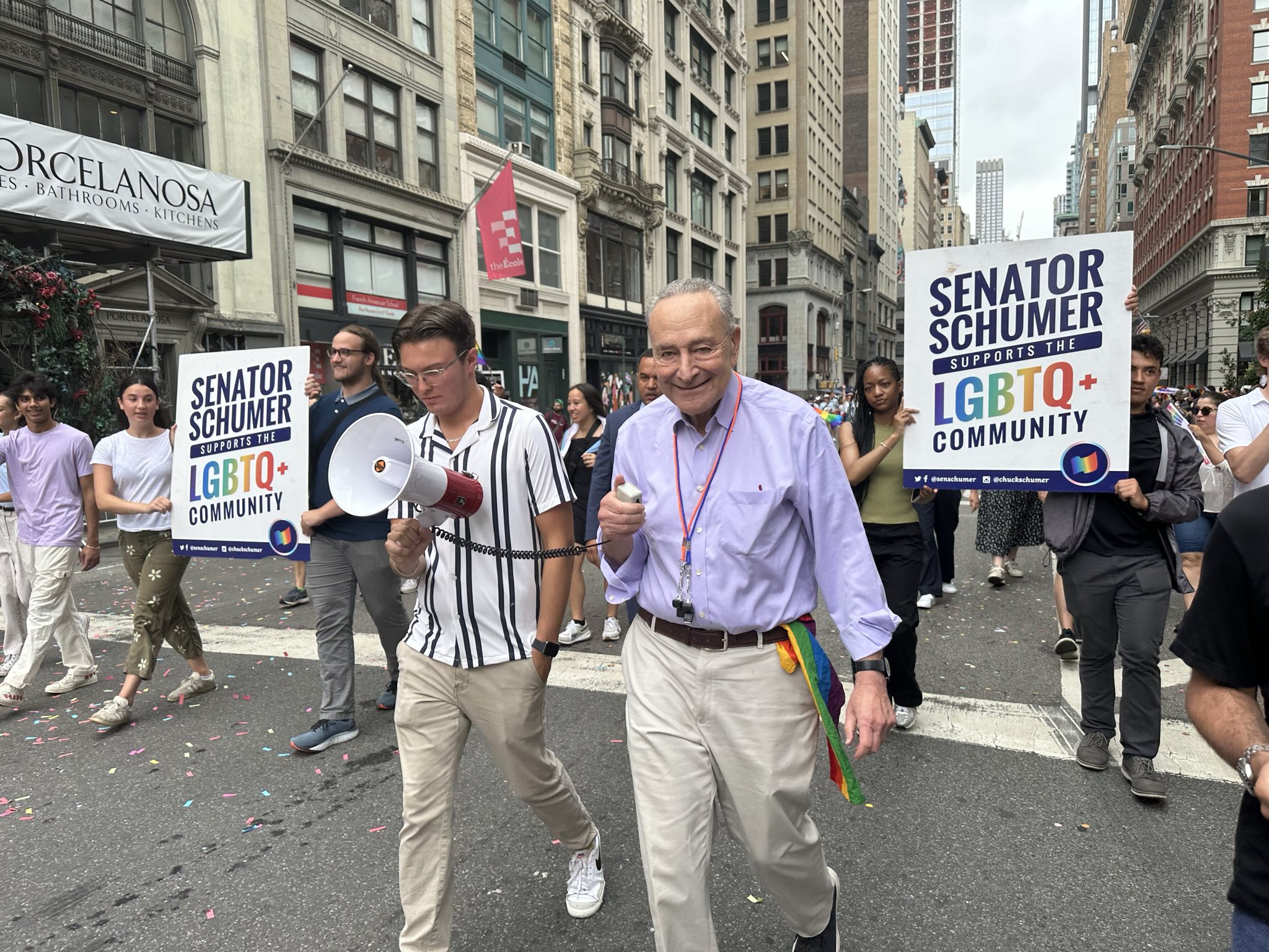 An old man walks with a crowd at a parade with people behind him holding several LGBTQ+ signs