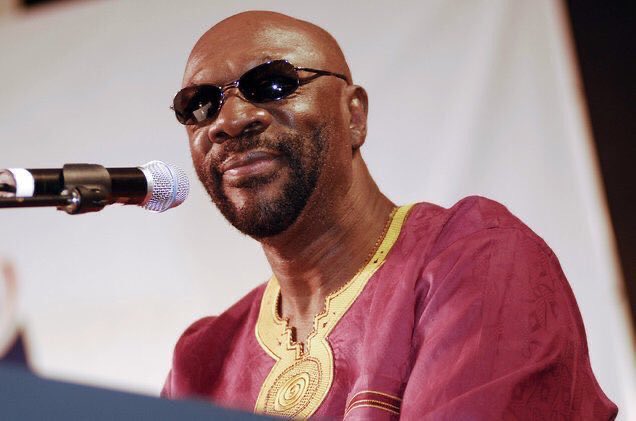 A man wearing a purple shirt and black sunglasses stands in front of a microphone