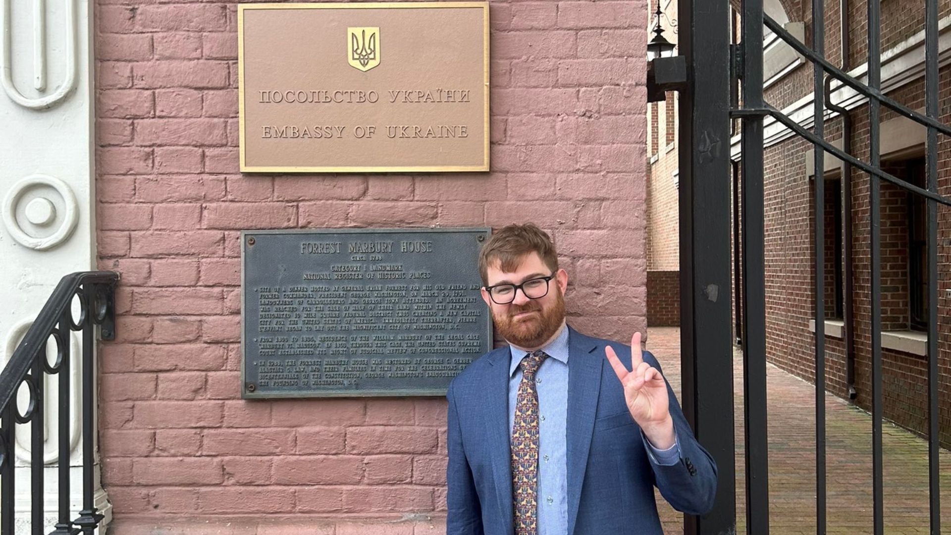 A man with red hair, glasses, and a suit standing next to the Embassy of Ukraine sign