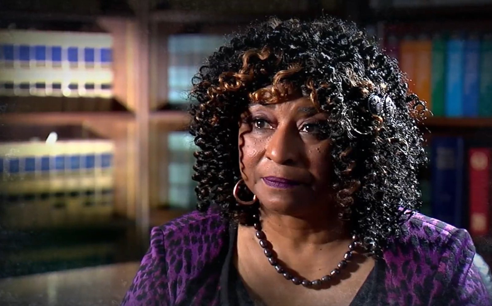 A woman with curly black hair wears a purple blouse while conducting an interview