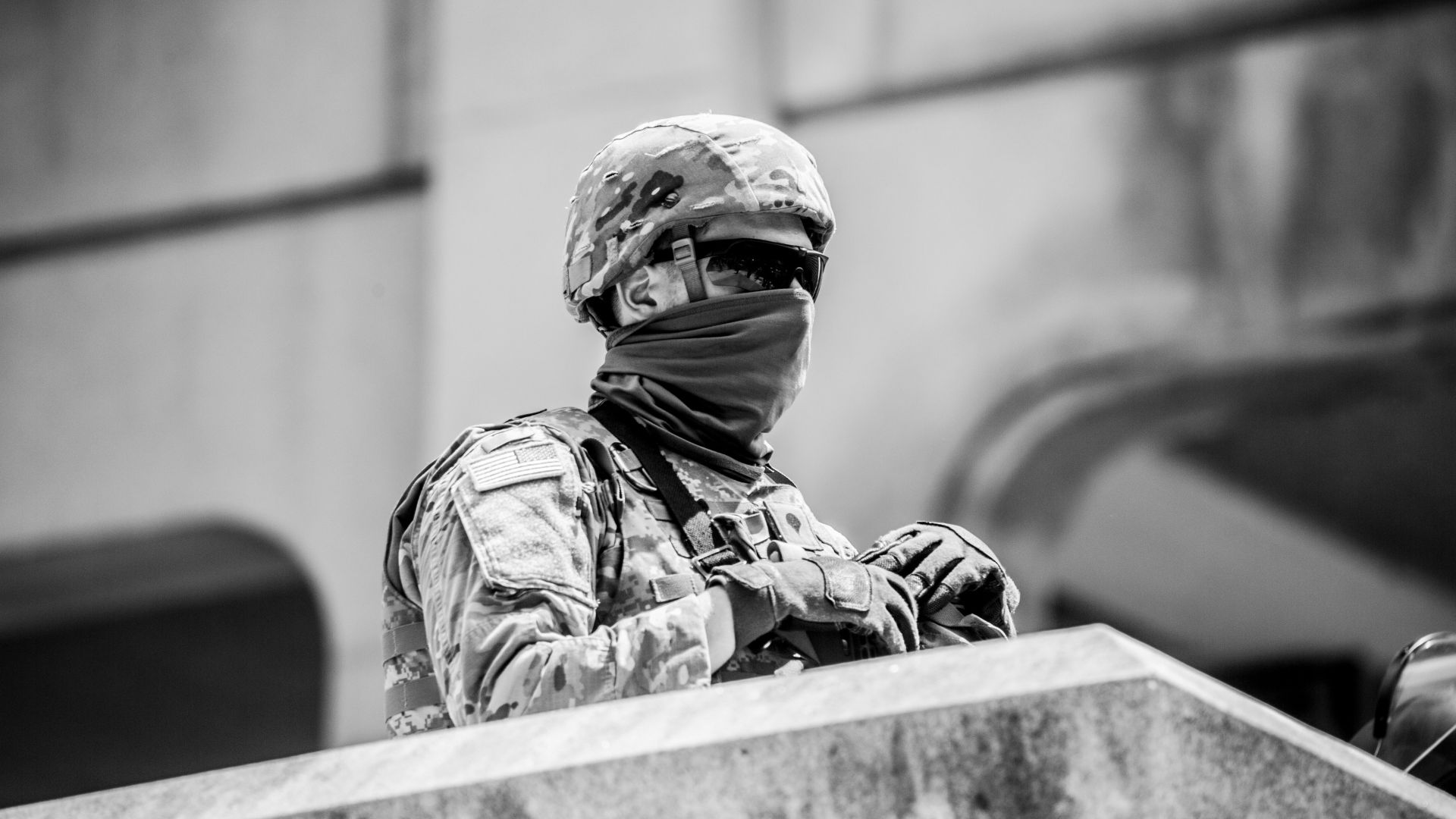 A man wearing army fatigues with a face mask, helmet, gloves, and sunglasses standing at the top of a set of stairs