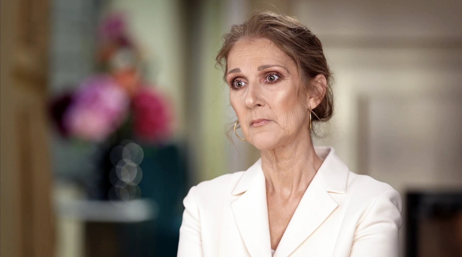 Celine Dion wears a white jacket and gold hoop earrings while sitting for an interview