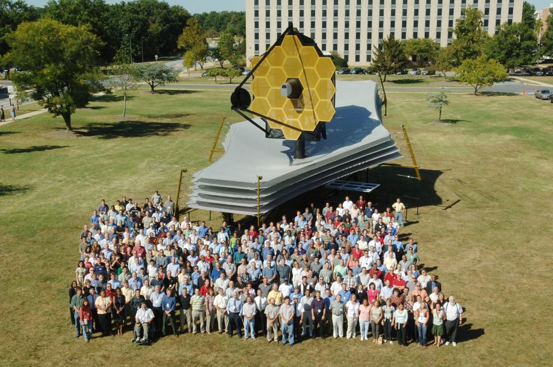 A photograph of a large group of researchers and scientists standing beside the James Webb Space Telescope