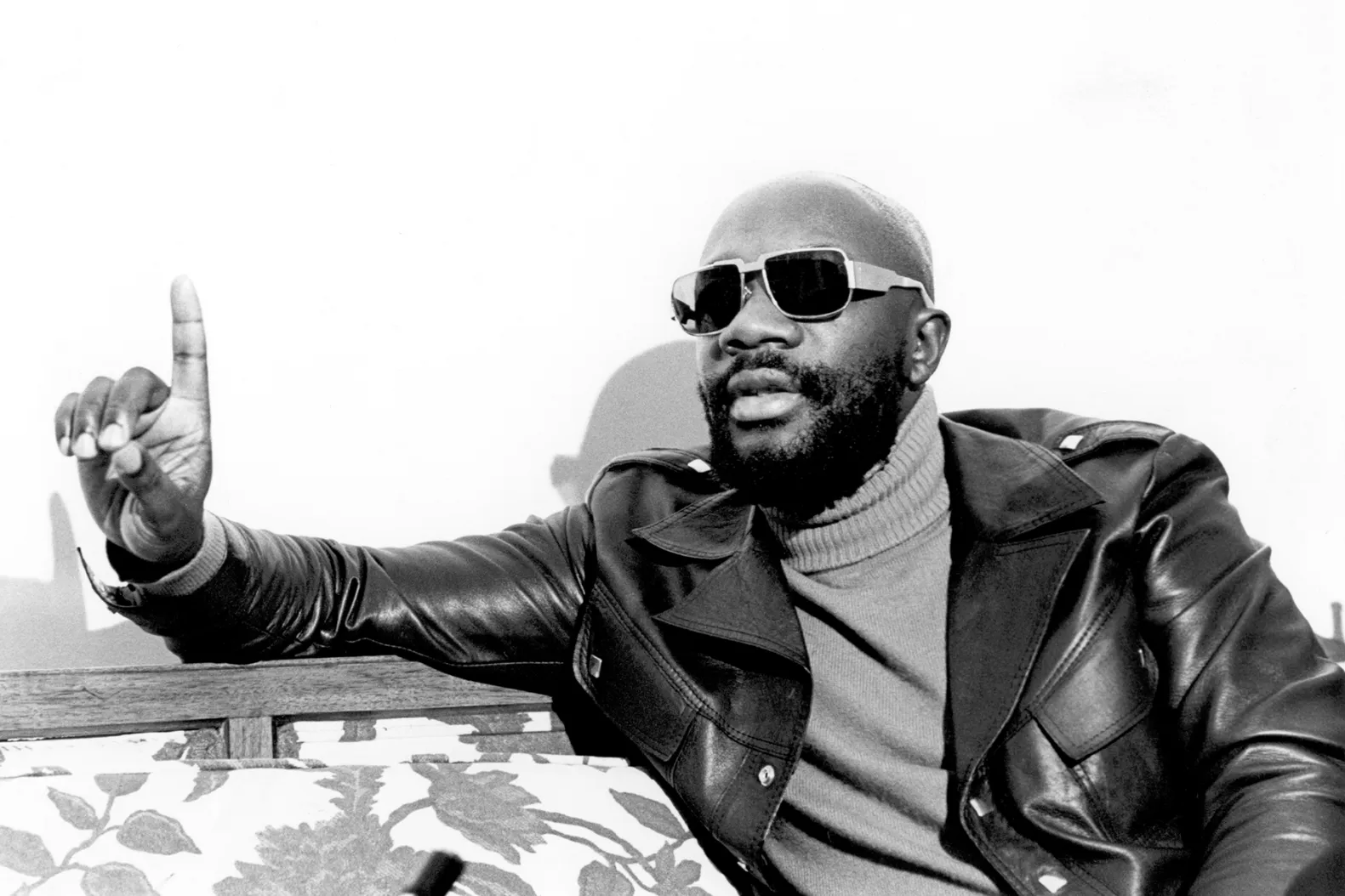 A man wearing a turtleneck sweater, leather coat, and sun glasses holds up his index finger