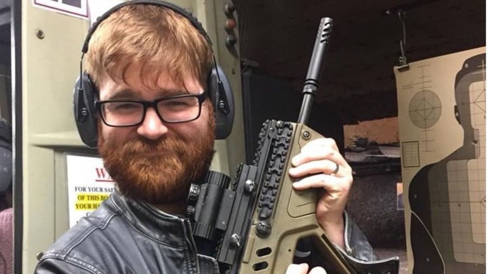 A man with red hair and glasses holding a machine gun