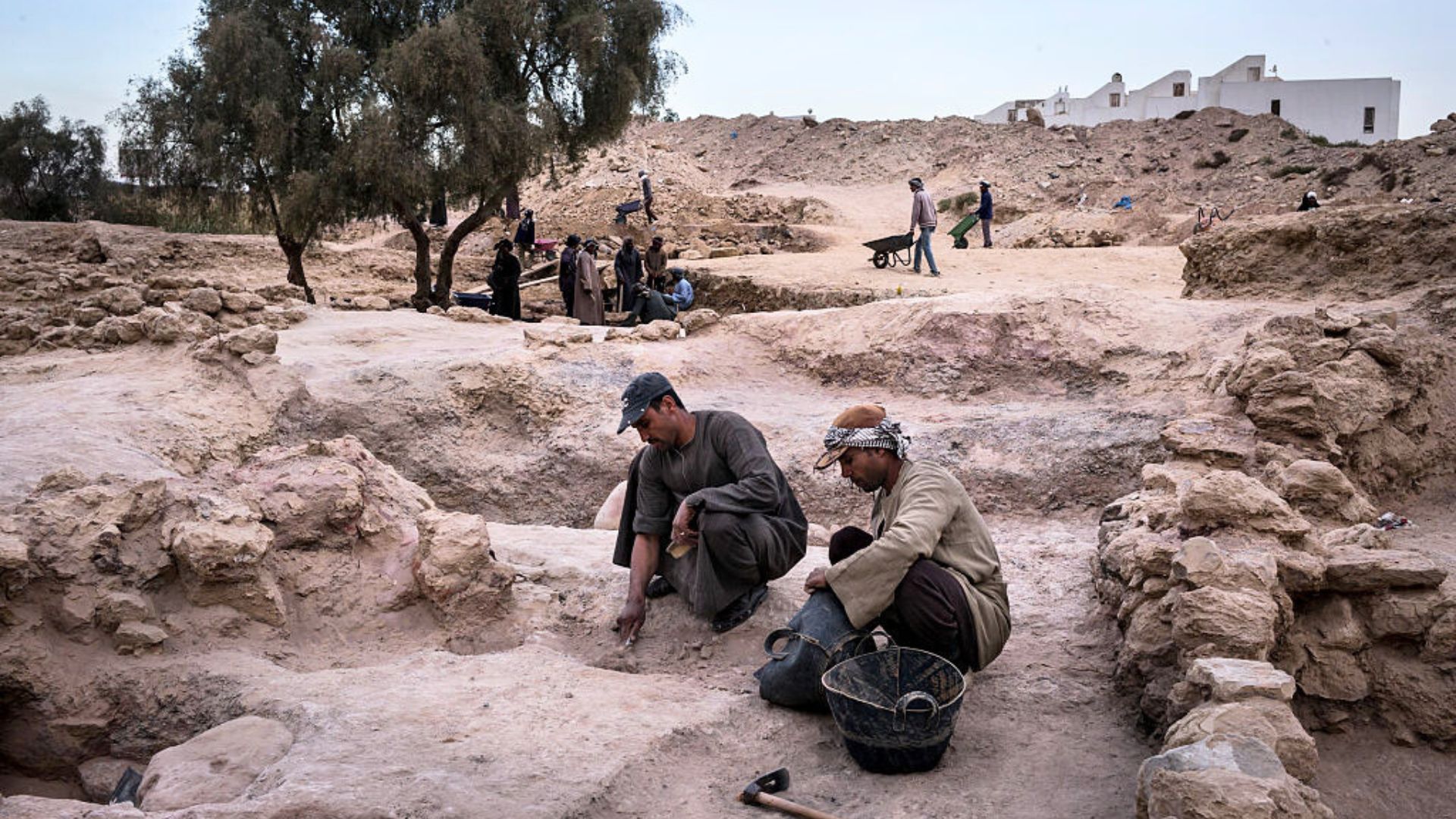 Two men crouch in a small archeological site in the desert surrounded by more sites and more men working in the hard ground