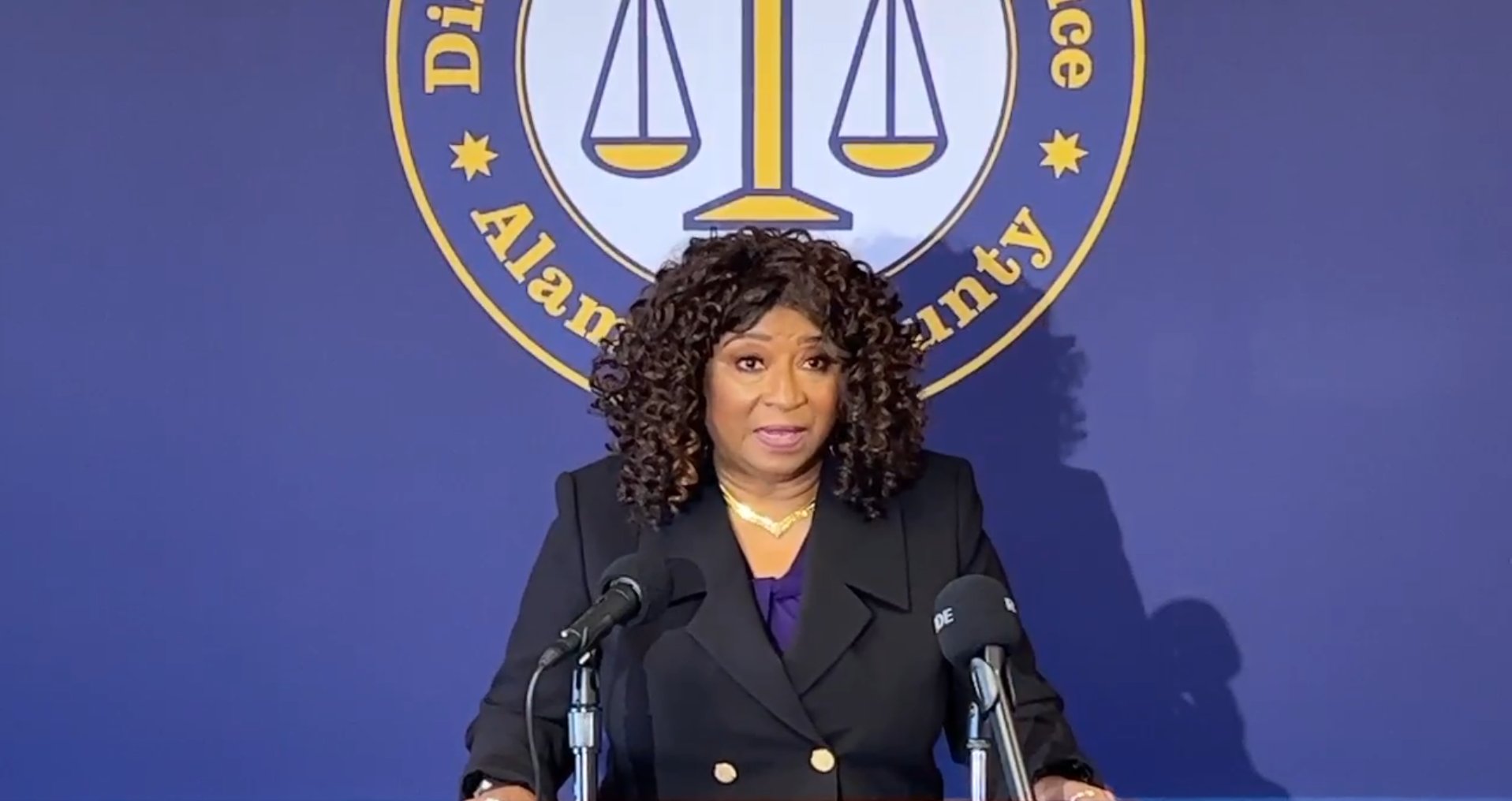 A woman with curly black hair stands at a set of microphones delivering a speech in front of an Alameda County sign